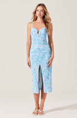 A woman is wearing a sleeveless, light blue dress with thin spaghetti straps. The dress has a keyhole cutout at the chest and features a watercolor-like pattern in various shades of blue. It is fitted at the waist and has a front slit that reveals part of her legs. She is also wearing white open-toe heels.