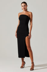 A woman is wearing a strapless black dress that is fitted at the top and extends to mid-calf length. The dress features a high slit on the left side, revealing her leg. She is accessorized with gold hoop earrings, a gold bracelet on her right wrist, and rings on her fingers. She is also wearing black high-heeled sandals with ankle straps.