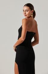 A woman is wearing a strapless, black dress with a fitted bodice that features visible seams and a hidden zipper on the side. The dress has a slit on the side of the skirt, revealing her leg. She is accessorized with large, gold hoop earrings and a thin gold bracelet on her wrist.