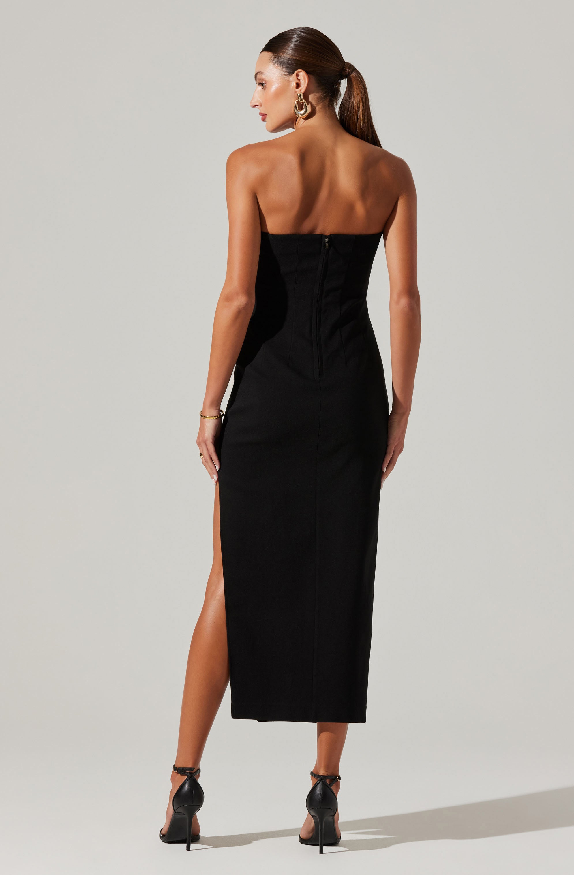 A woman is wearing a strapless, black, full-length dress with a slit on one side. The dress has a fitted silhouette with a back zipper closure. She is also wearing high-heeled sandals and gold jewelry, including hoop earrings, a bracelet, and rings. Her hair is styled in a sleek, low ponytail.