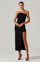 A woman is wearing a strapless, form-fitting black dress that has a high slit on the left side, revealing her leg. She is accessorized with gold earrings, a gold bracelet, and rings. She is wearing black high-heeled sandals with ankle straps.