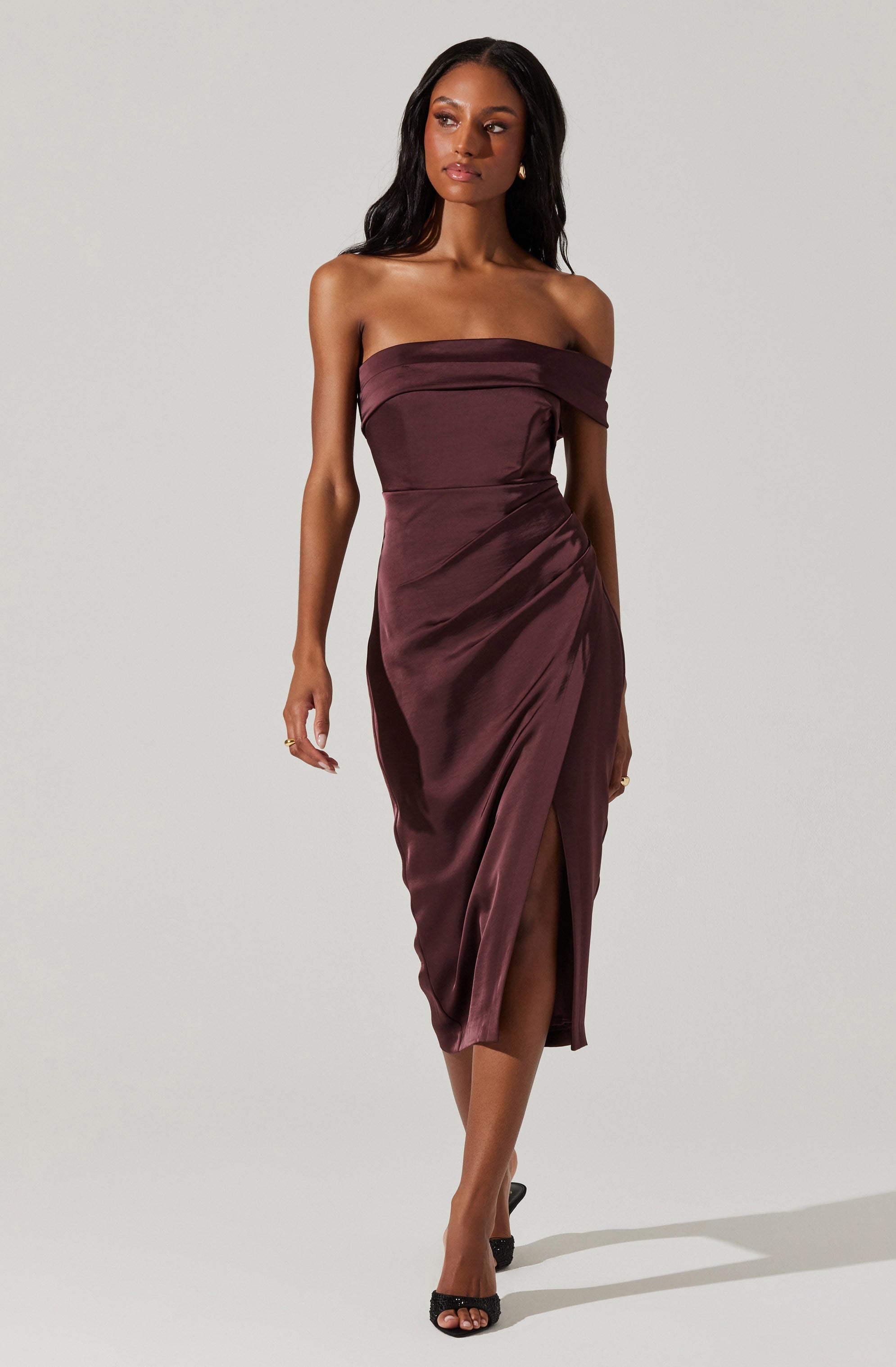 A woman is wearing a strapless, off-the-shoulder, dark maroon satin dress with a fitted bodice and a side slit that reveals her leg. The dress features a draped detail at the top and gathers at the waist, creating a sleek and elegant silhouette. She is also wearing black high-heeled sandals.