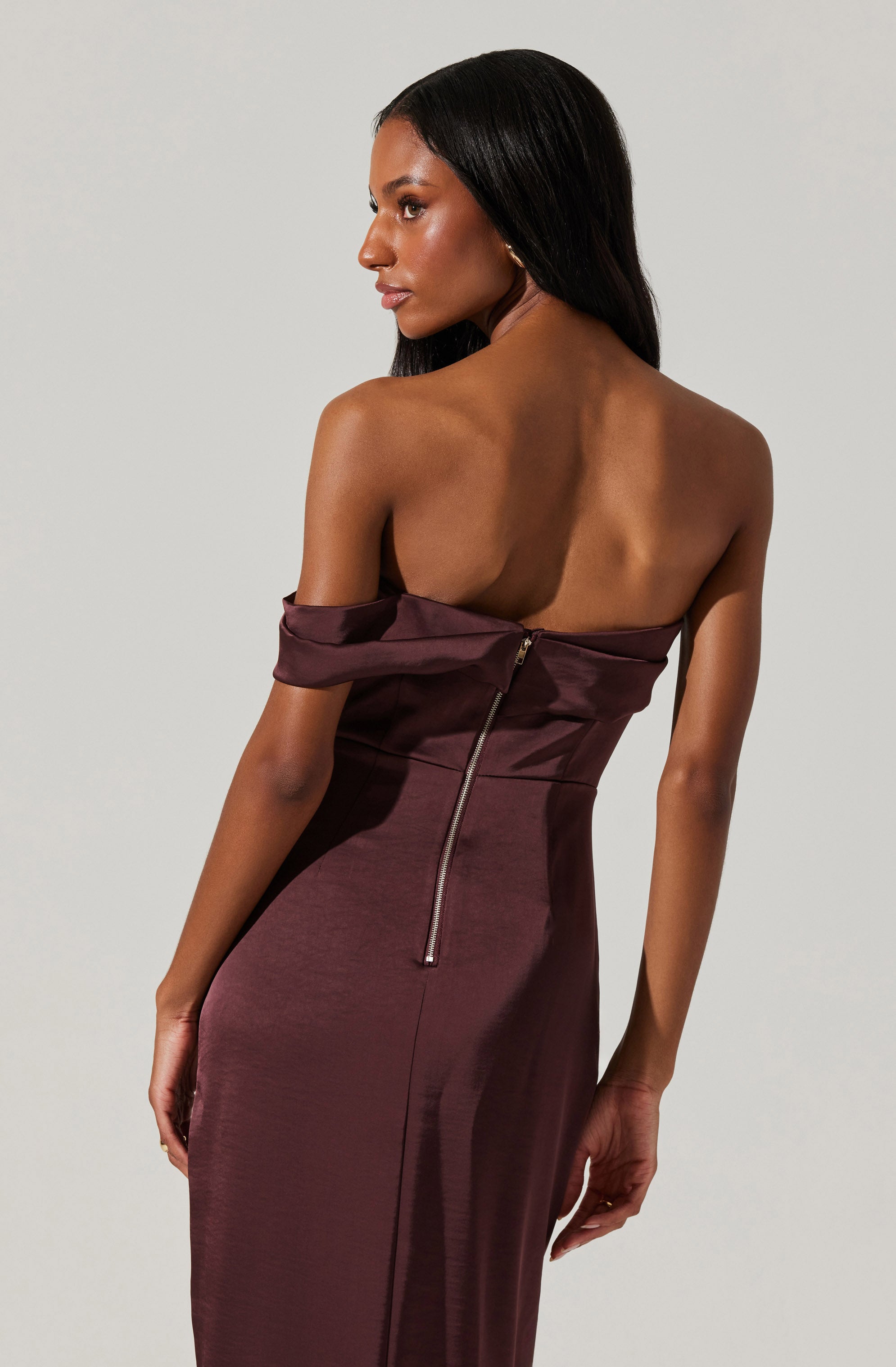 A woman is wearing an elegant, strapless, deep burgundy satin dress with a fitted silhouette. The dress features a visible gold zipper running down the back, which is partially unzipped at the top, creating an off-the-shoulder effect on one side. The fabric has a smooth, shiny finish, and the dress drapes gracefully along her body.