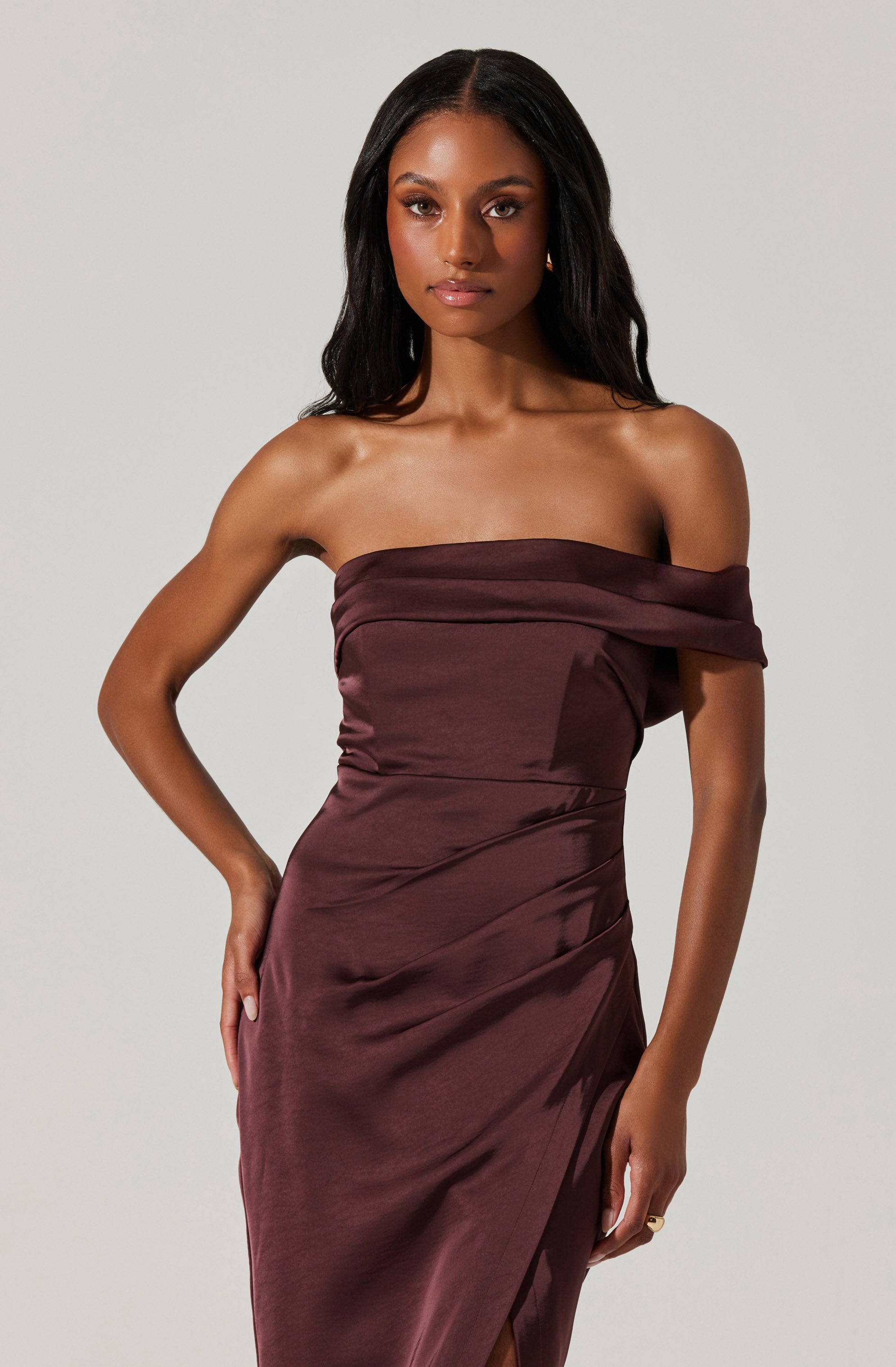 A woman is wearing a strapless, off-the-shoulder dress in a deep burgundy color. The dress features a ruched bodice with a wide, folded neckline that drapes around her shoulders. The fabric appears smooth and shiny, with a slit on one side.