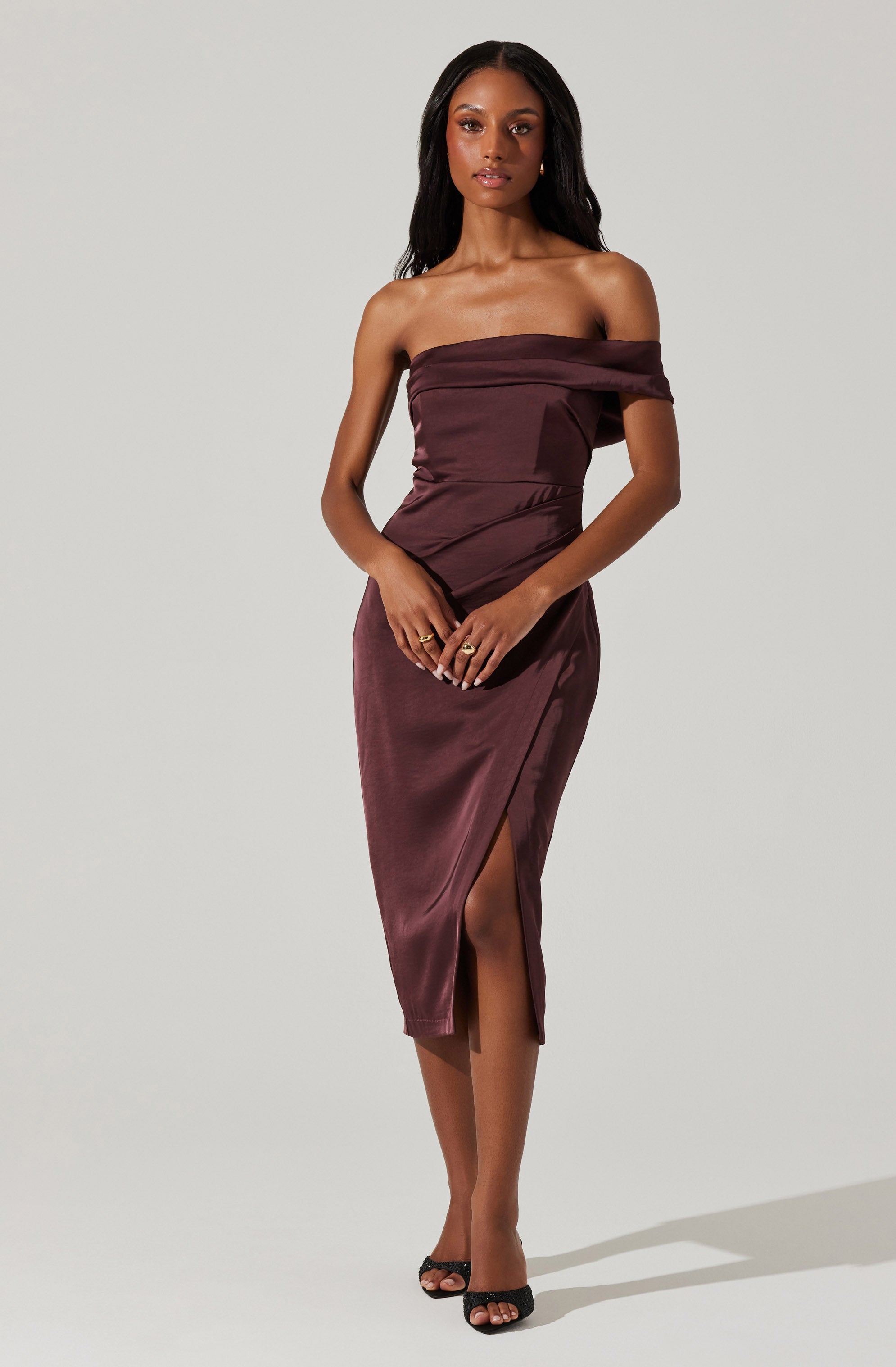 A woman is wearing an off-the-shoulder, deep burgundy satin dress that is fitted at the waist and features a high slit on the front left side. The dress has a draped, ruched detail across the bodice. She is also wearing black open-toe heels.