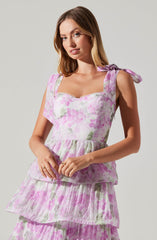A woman is wearing a sleeveless, tiered dress with a pink and white floral pattern. The dress has shoulder straps tied into bows on each shoulder. The fabric appears lightweight and textured, with multiple ruffled layers creating a flowing, layered look.