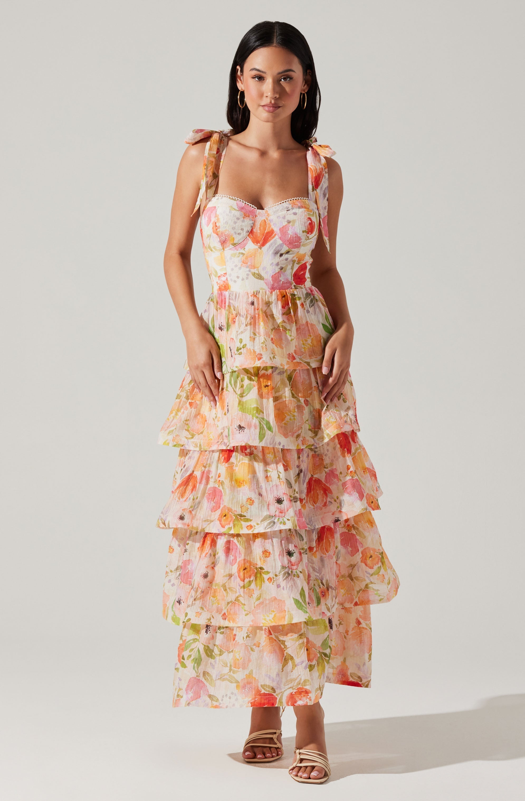 A woman is wearing a sleeveless, tiered maxi dress with shoulder straps tied into bows. The dress features a light, airy fabric with a colorful floral pattern in shades of pink, orange, yellow, and green. She completes her look with a pair of open-toe, strappy sandals in a neutral tone and gold hoop earrings.