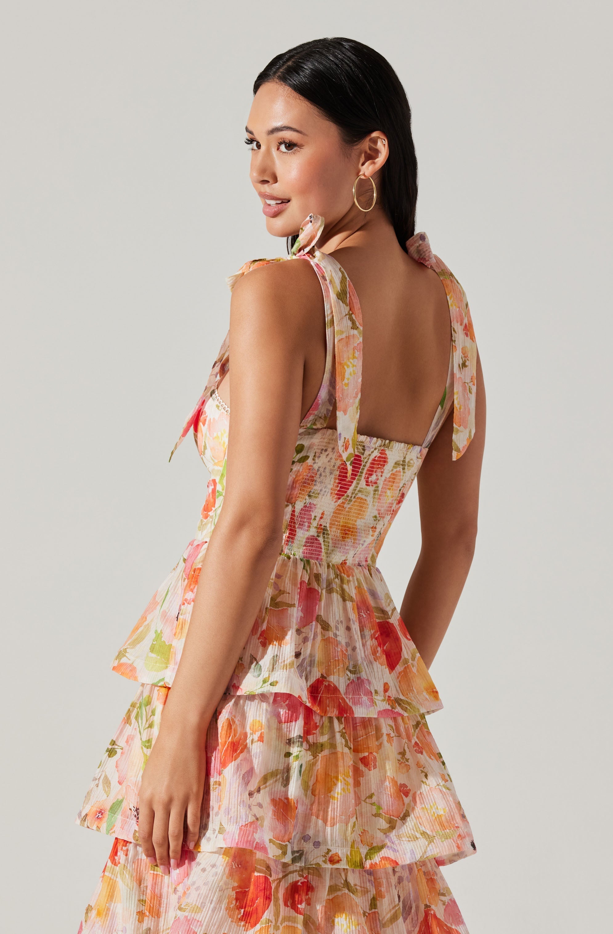 A woman is wearing a sleeveless, floral-print dress with a fitted bodice and layered, ruffled skirt. The dress features shoulder straps tied into bows, and the fabric has a light, airy texture with a colorful pattern of pink, orange, green, and cream hues.
