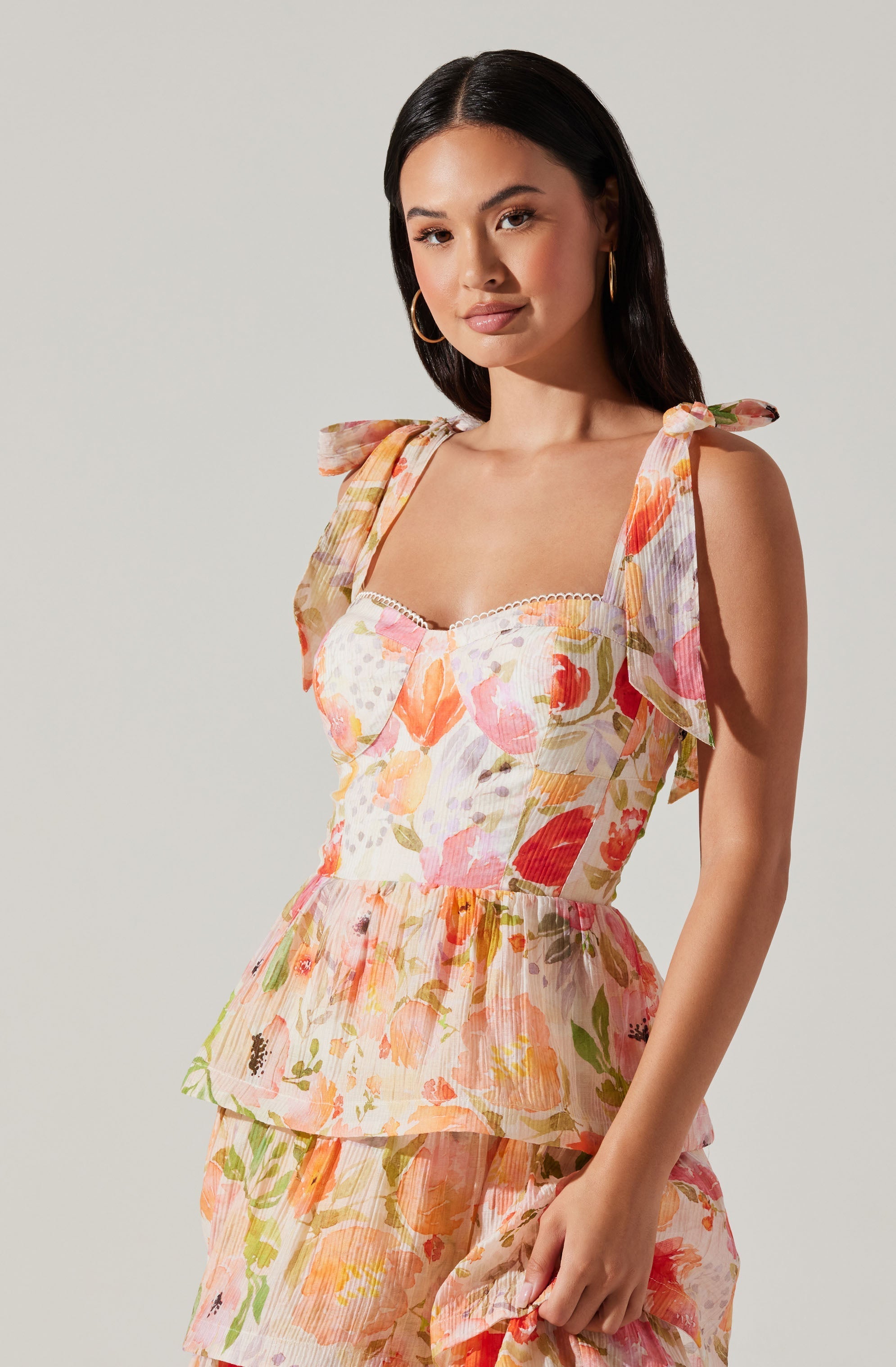 A woman is wearing a sleeveless, floral-patterned dress with ruffled shoulder straps tied into bows. The dress features a fitted bodice with a subtle sweetheart neckline and a tiered skirt. The fabric has a light, airy texture with a colorful print of pink, orange, yellow, and green flowers and leaves.