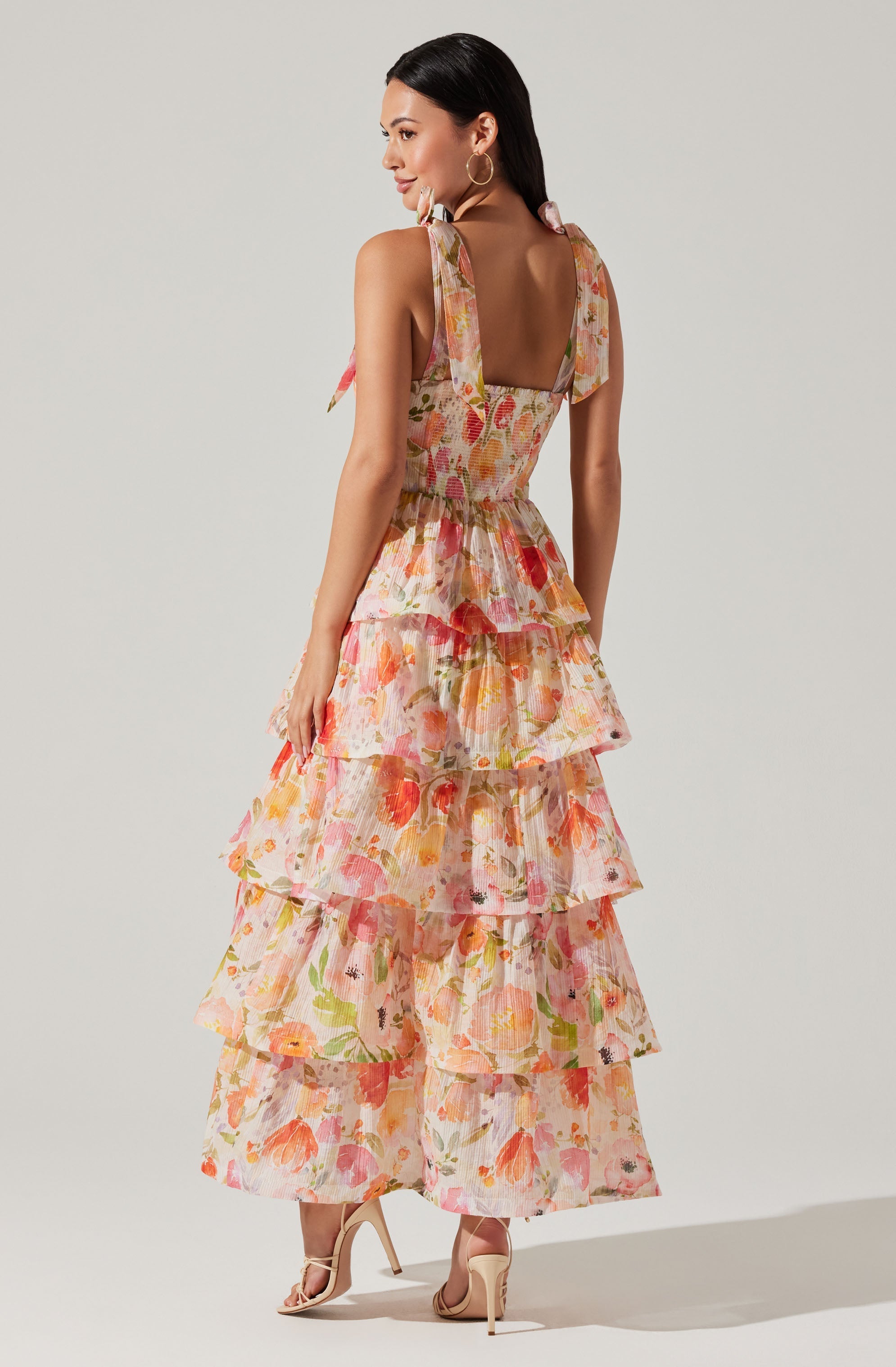 A woman is wearing a sleeveless, tiered maxi dress with a floral print in shades of pink, orange, yellow, and green. The dress features shoulder straps tied into bows on each shoulder, a fitted bodice, and a flowing skirt with multiple ruffled layers. She is also wearing nude high-heeled sandals and large hoop earrings.