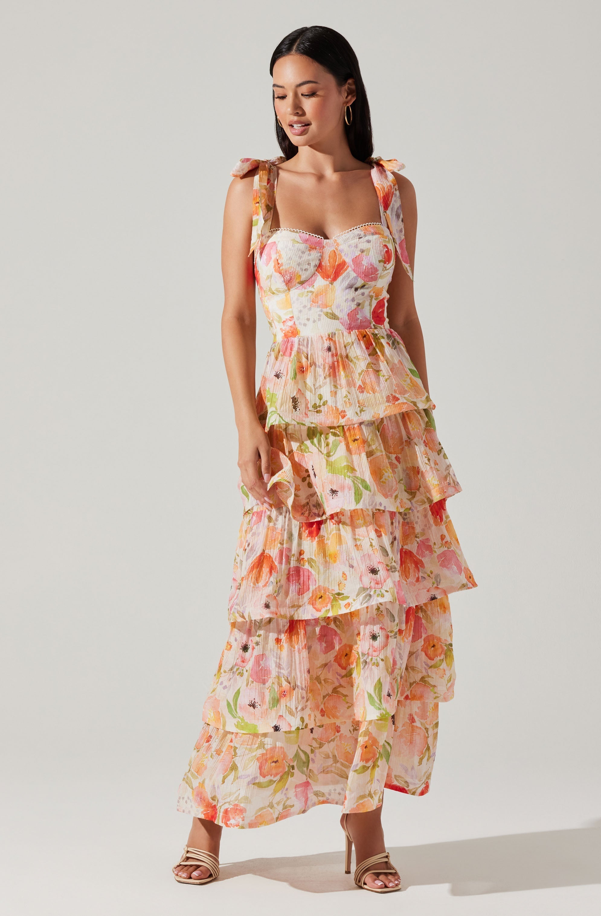 A woman is wearing a sleeveless, tiered maxi dress with shoulder ties. The dress features a colorful, floral pattern with shades of pink, orange, green, and cream. The bodice has a sweetheart neckline with a lace trim detail along the top edge. She is also wearing open-toe high-heeled sandals.