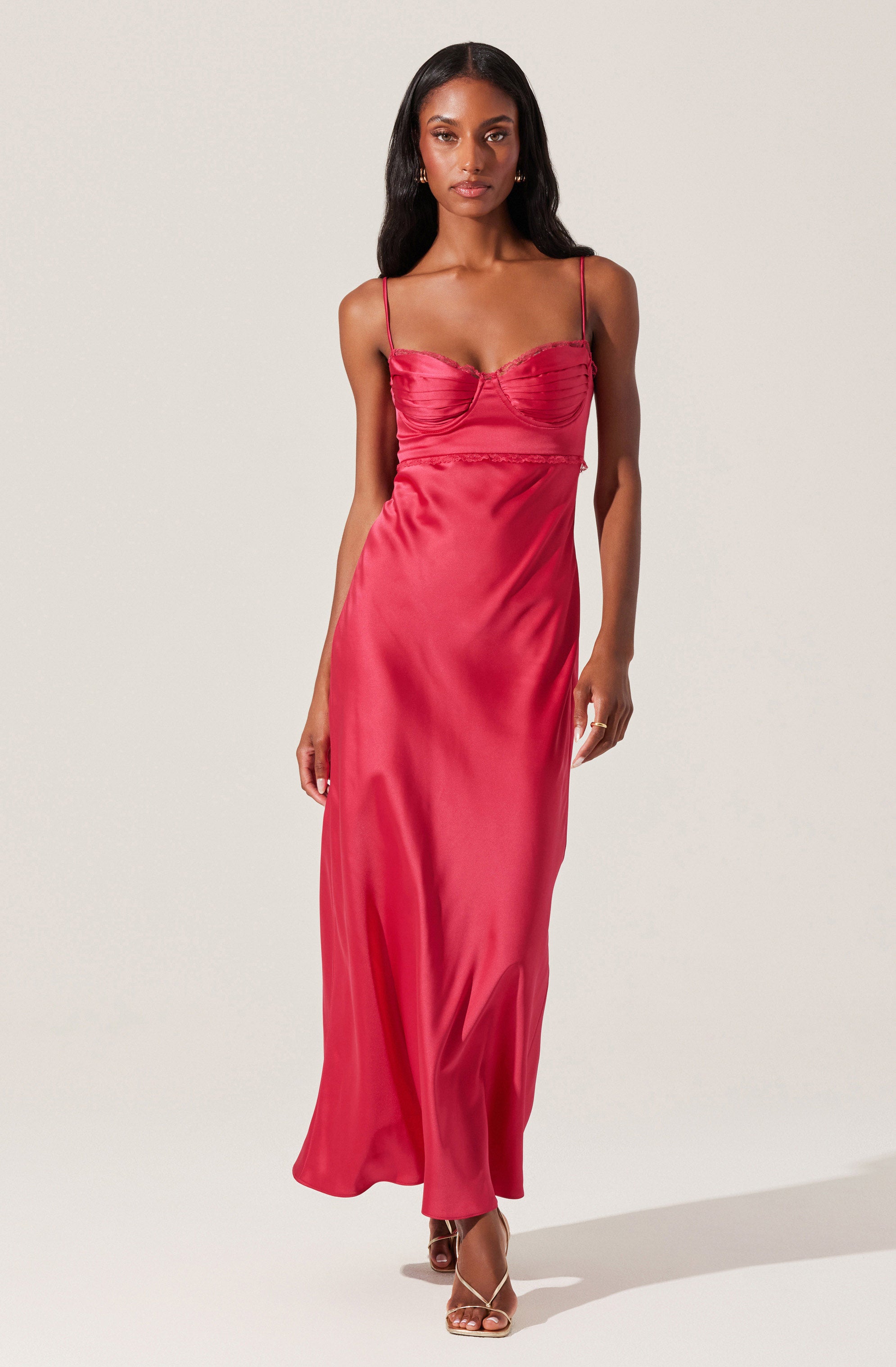 A woman is wearing a long, satin, spaghetti-strap dress in a deep pink color. The dress has a fitted bodice with ruched detailing at the bust and a smooth, flowing skirt that reaches the floor. The thin straps sit on her shoulders, and she is wearing open-toe heels.
