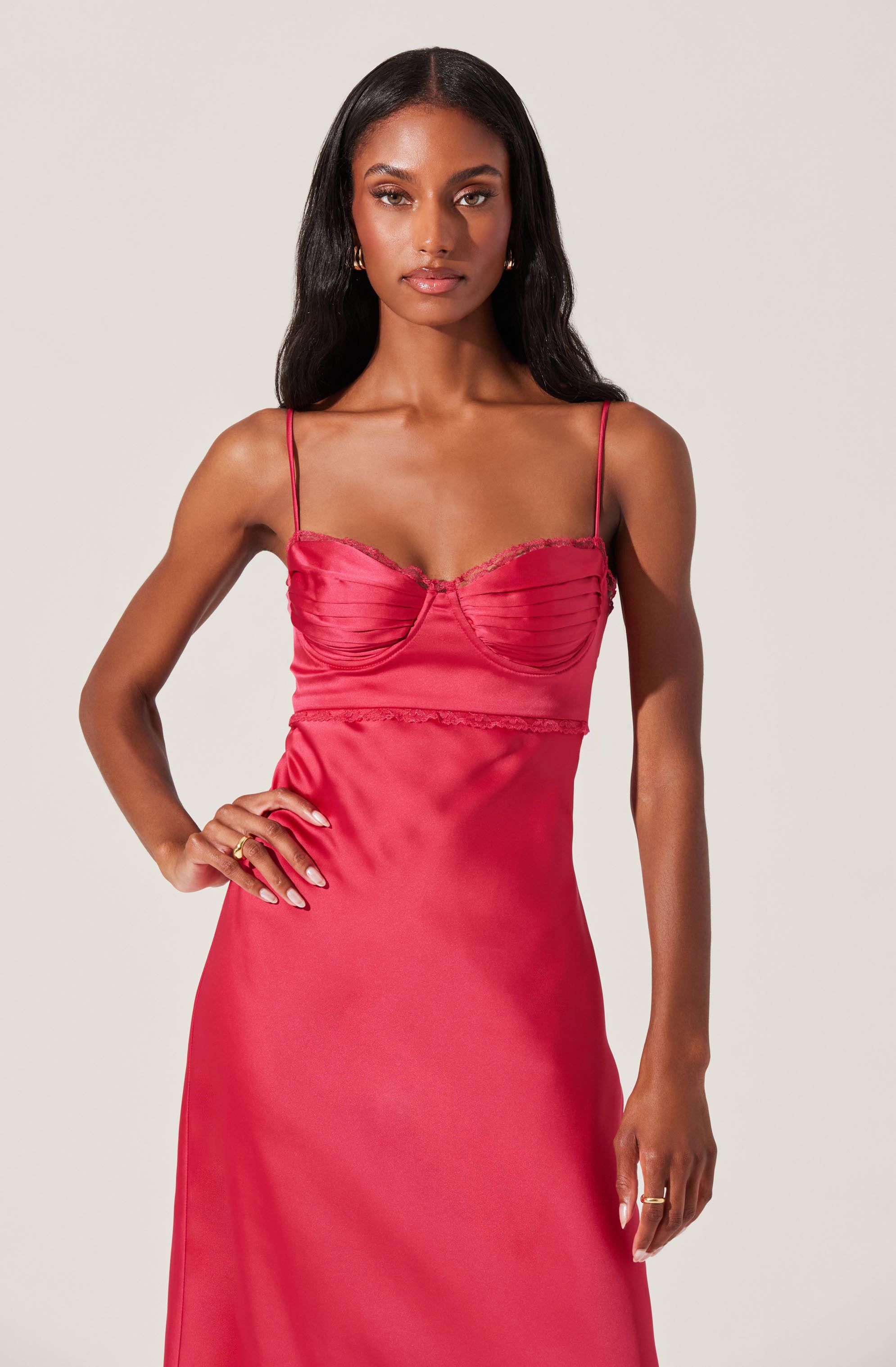 A woman is wearing a sleeveless, slip-style satin dress in a bright pink color. The dress has thin spaghetti straps, a ruched bodice with a sweetheart neckline, and lace trim details at the bust and waist.