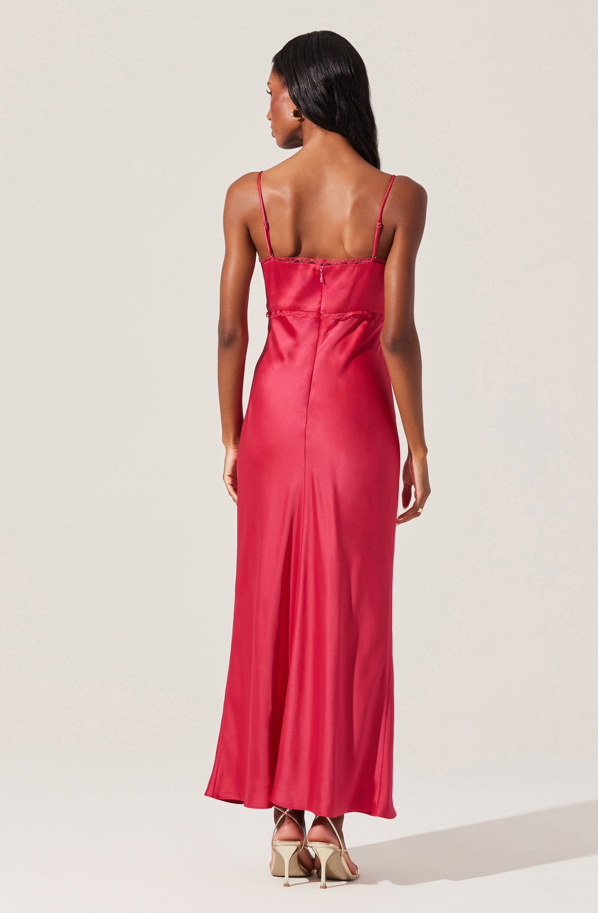 A woman is wearing a long, elegant, satin slip dress in a vibrant shade of pink. The dress features thin spaghetti straps and a fitted bodice with a slight ruffle detail along the top edge. The fabric of the dress is smooth and shiny, flowing down to the floor with a gentle flare at the hem. She is wearing high-heeled shoes with ankle straps.