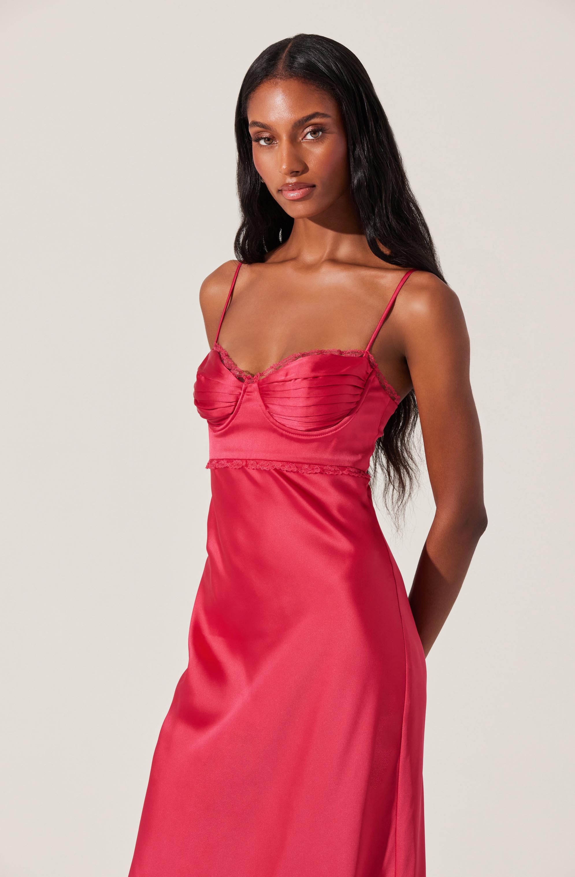 A woman wearing a silky, deep pink slip dress with spaghetti straps, featuring a ruched bust detail with lace trim. The dress is fitted at the top and flows loosely down to the hem.