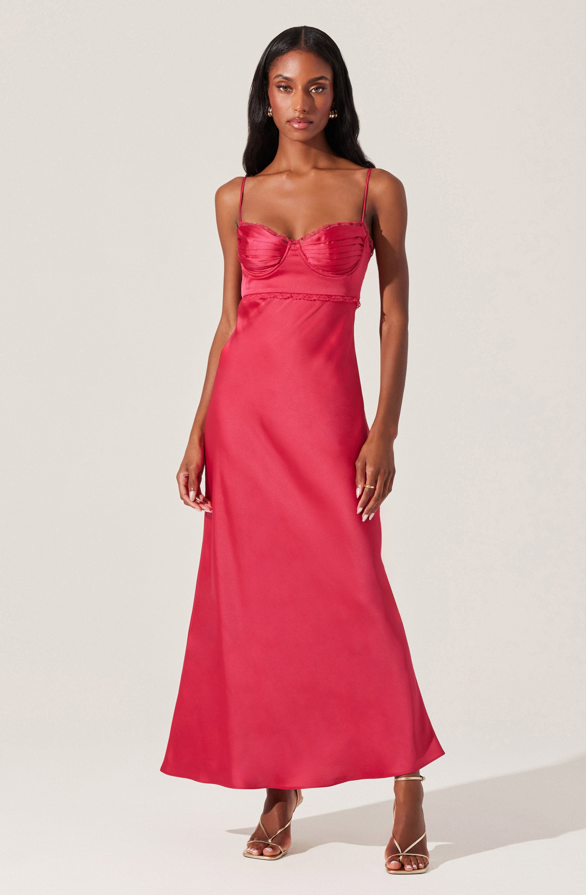 A woman wearing a floor-length, sleeveless, spaghetti strap dress in a vibrant pink color. The dress features a fitted bodice with a ruched detail across the bust and a slight empire waistline. The skirt flows smoothly down to the floor, creating an elegant silhouette. She pairs the dress with strappy high-heeled sandals.