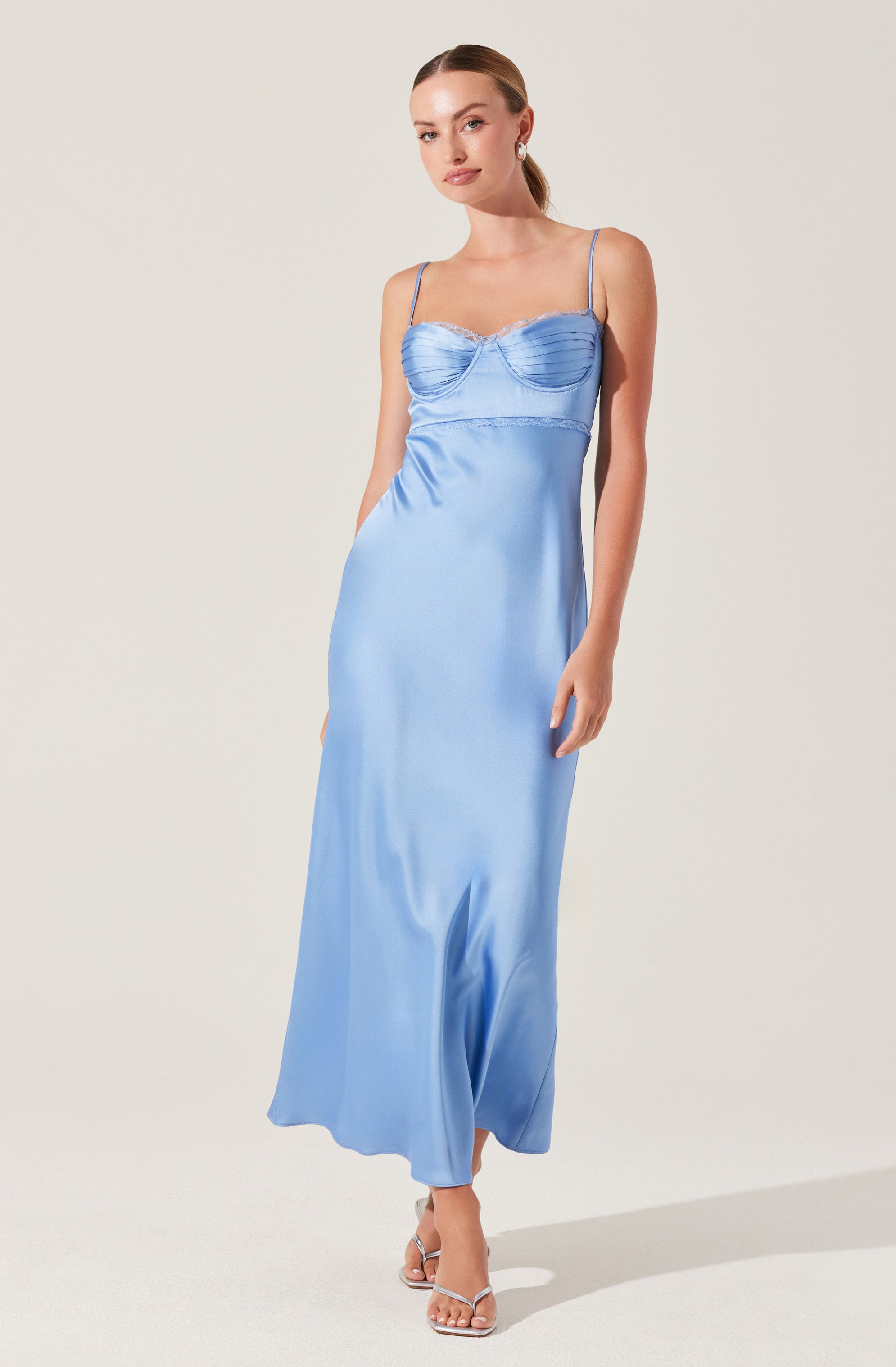 A woman is wearing a full-length, light blue satin dress with thin spaghetti straps. The bodice features ruched detailing and a subtle sweetheart neckline, with a fitted waist flowing into a smooth, flowing skirt that reaches the floor. She is also wearing clear strappy high-heeled sandals.