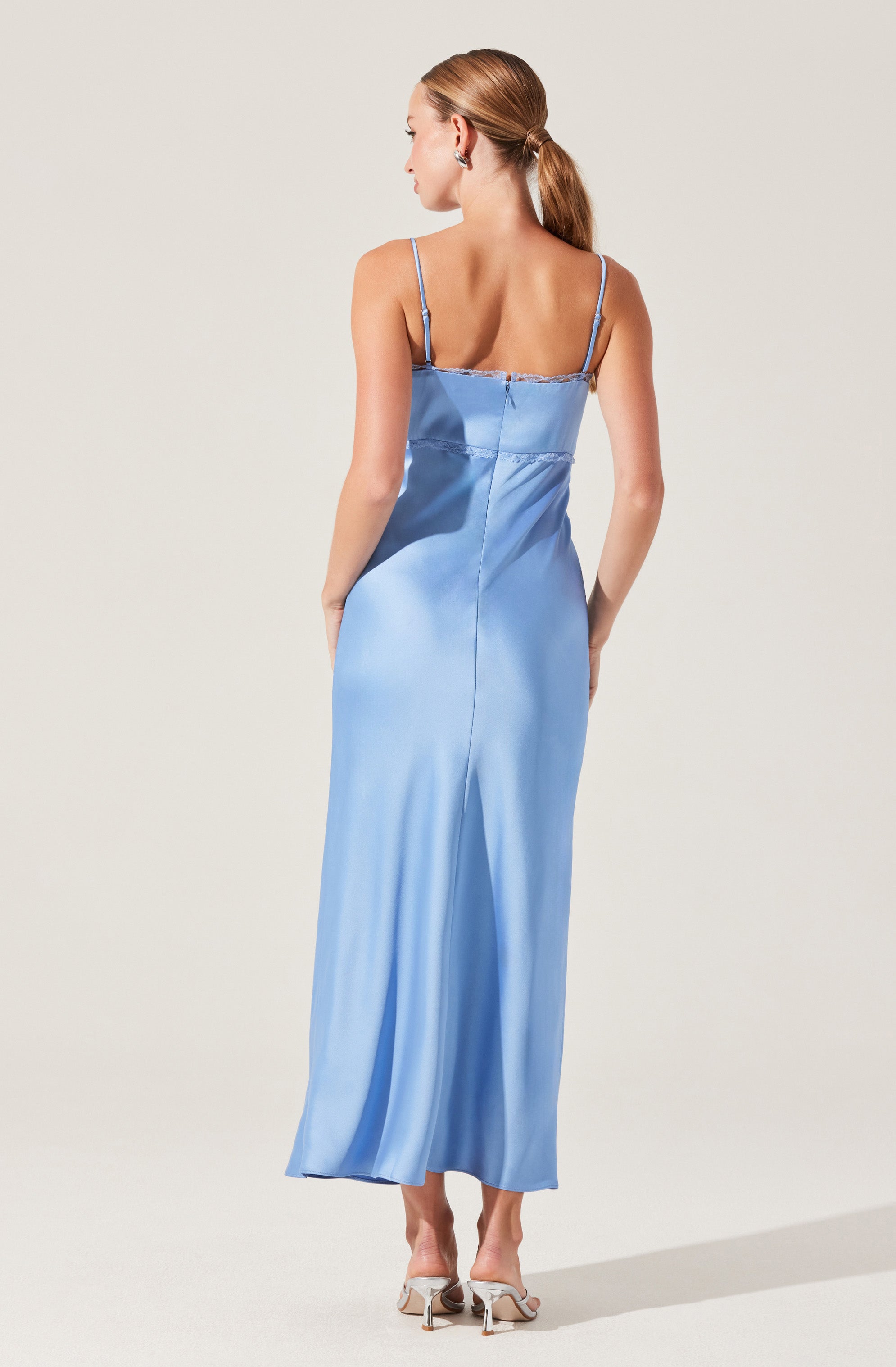 A woman is wearing a light blue satin slip dress with thin spaghetti straps. The dress features a straight neckline with a delicate lace trim. It has a fitted bodice and flows into a long, loose skirt that reaches the floor. The back of the dress is simple with a center seam running from the top to the hem. She completes her look with nude high-heeled sandals.