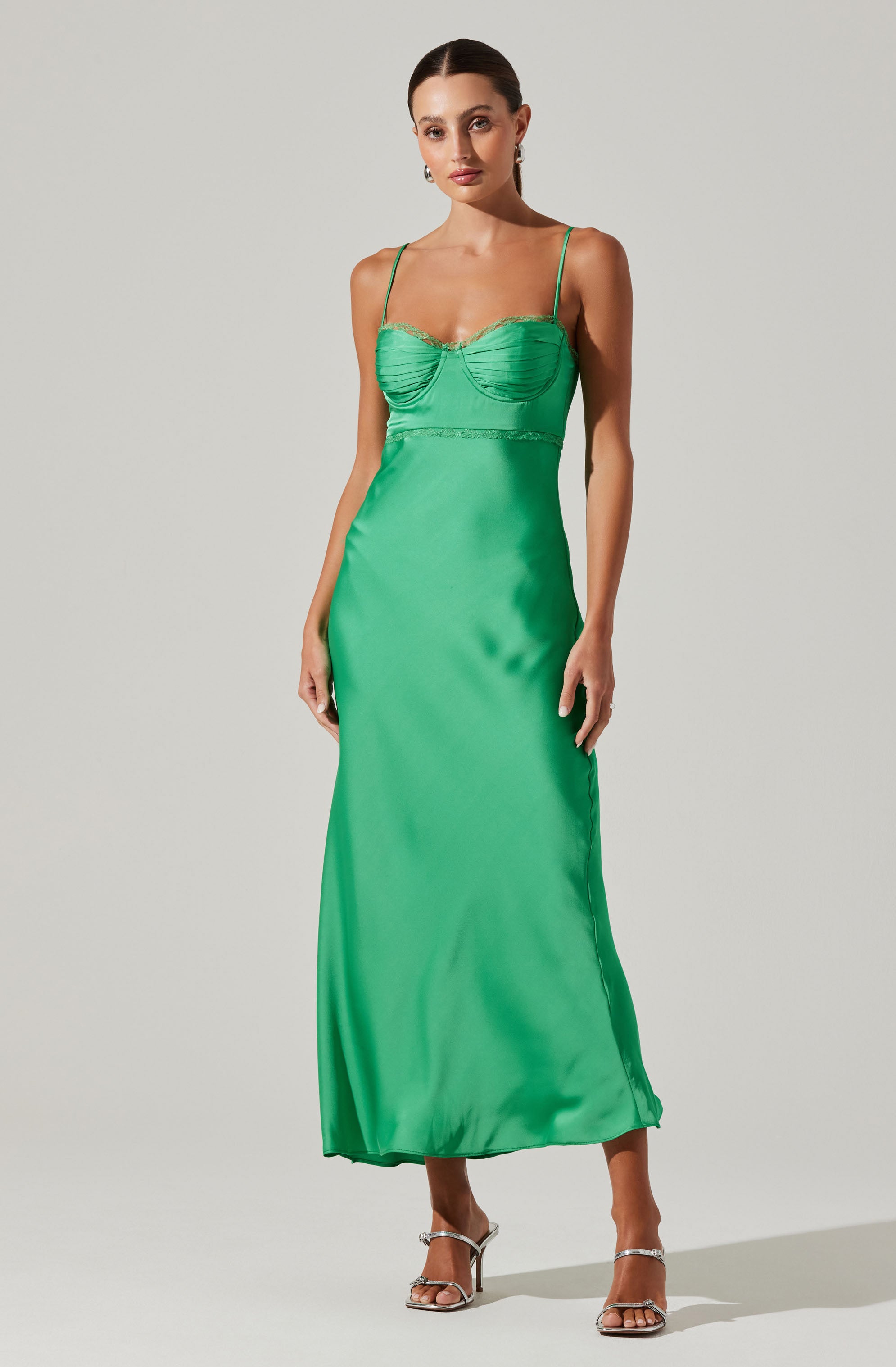 A woman is wearing a long, flowing, emerald green satin dress with thin spaghetti straps. The dress features a fitted bodice with ruching details and a fitted waist, flowing down to a slightly flared hem. She has paired the dress with open-toed, high-heeled sandals.
