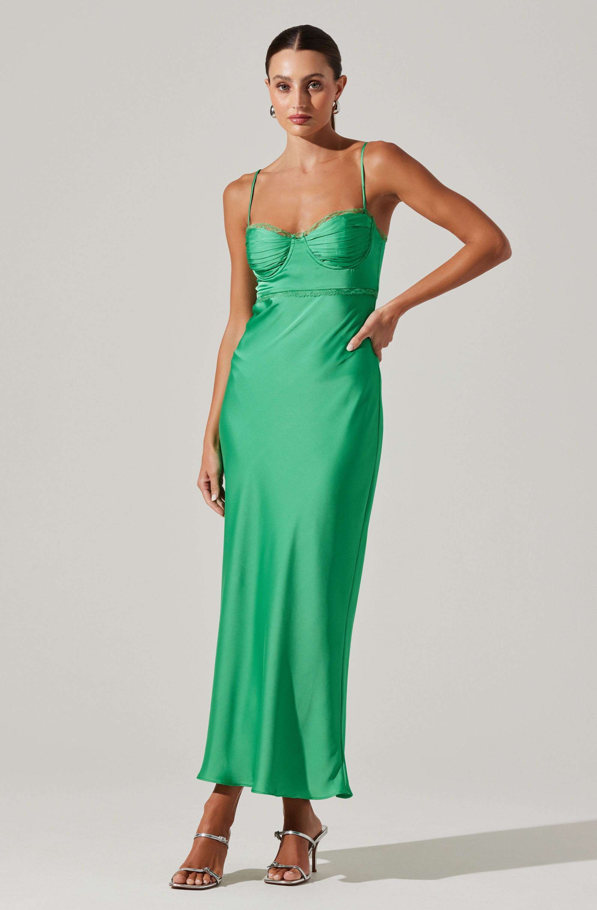 A woman is wearing a long, bright green satin dress with thin spaghetti straps. The dress has a fitted bodice with ruching detail and a smooth, flowing skirt that reaches the floor. She is also wearing silver high-heeled sandals.