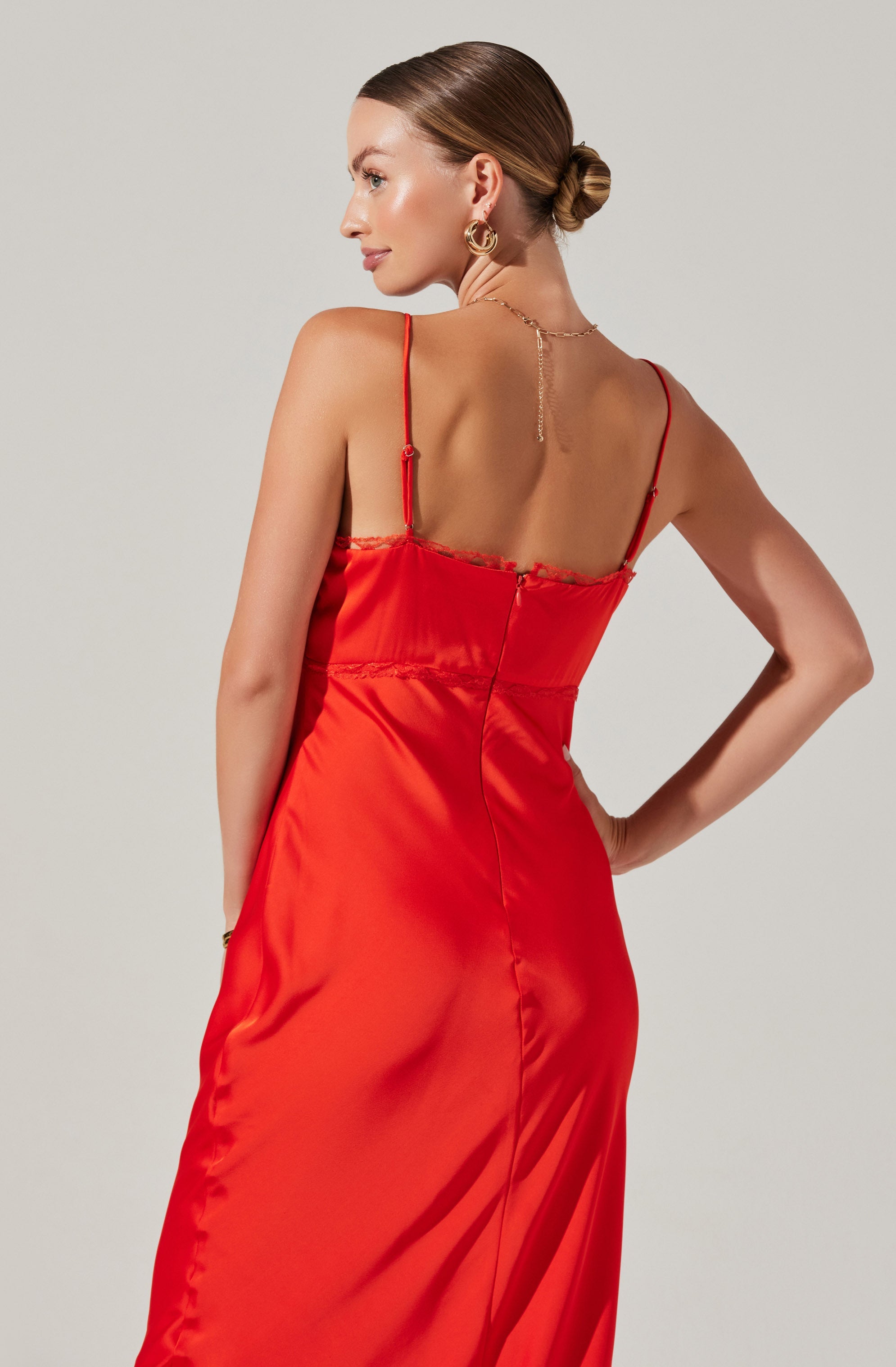A woman wearing a long, silky red dress with thin spaghetti straps and a fitted bodice. The dress features delicate lace trim along the top edge and a central back zipper. She accessorizes with gold hoop earrings, a gold chain necklace, and has her hair styled in a low bun.