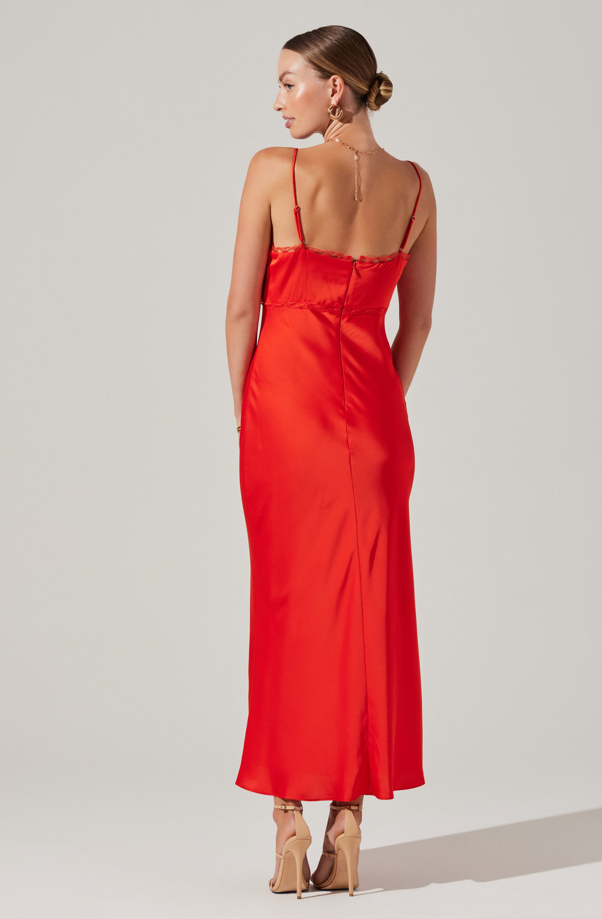 A woman is wearing a long, red satin slip dress with thin spaghetti straps. The dress features a gathered detail around the bust and a center back zipper. She has paired the dress with nude high-heeled shoes and is accessorized with gold jewelry, including hoop earrings and a delicate necklace.