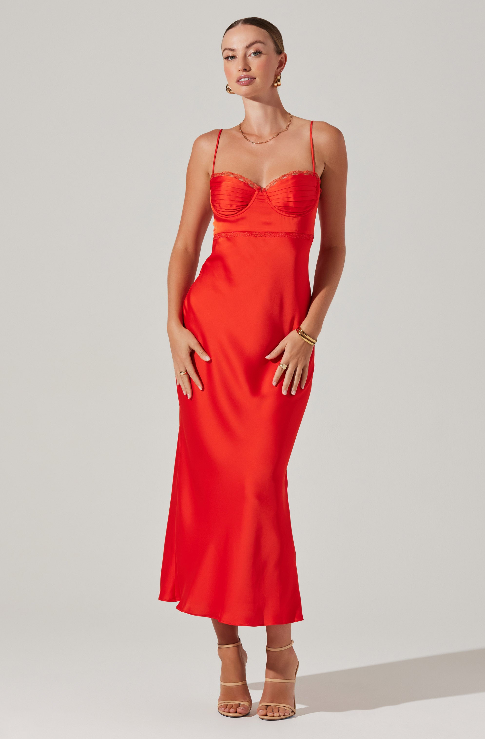 A woman is wearing a long, fitted red dress with thin spaghetti straps. The dress features a ruched bust area and a smooth, flowing skirt that reaches down to her ankles. She accessorizes with gold hoop earrings, a delicate gold necklace, and gold bangles on her right wrist. She is also wearing beige strappy high-heeled sandals.