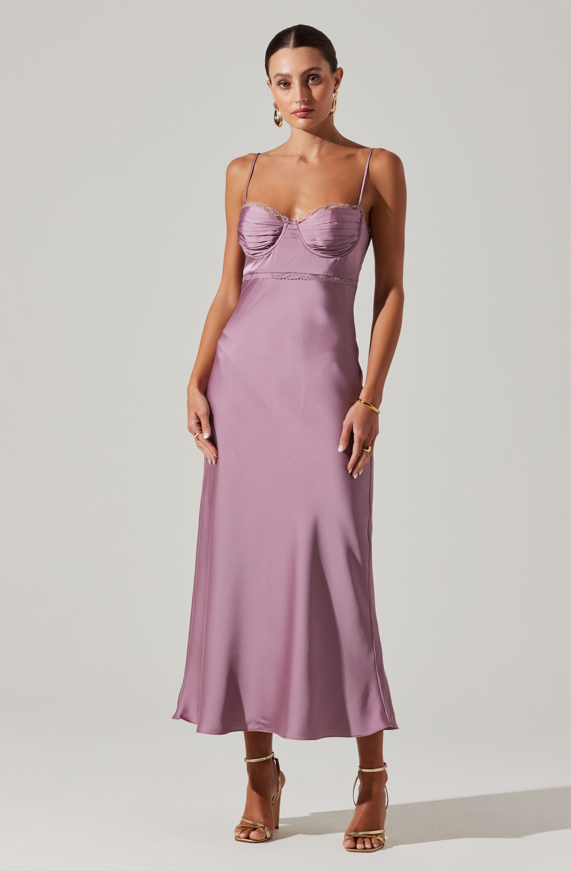 A woman is wearing a long, elegant lavender satin evening gown with thin spaghetti straps. The bodice features ruched detailing and a sweetheart neckline with lace trim. The dress has a fitted waist and flows down to a slightly flared hem, reaching just above her ankles. She is also wearing beige strappy high-heeled sandals and jewelry, including earrings, rings, and a bracelet.