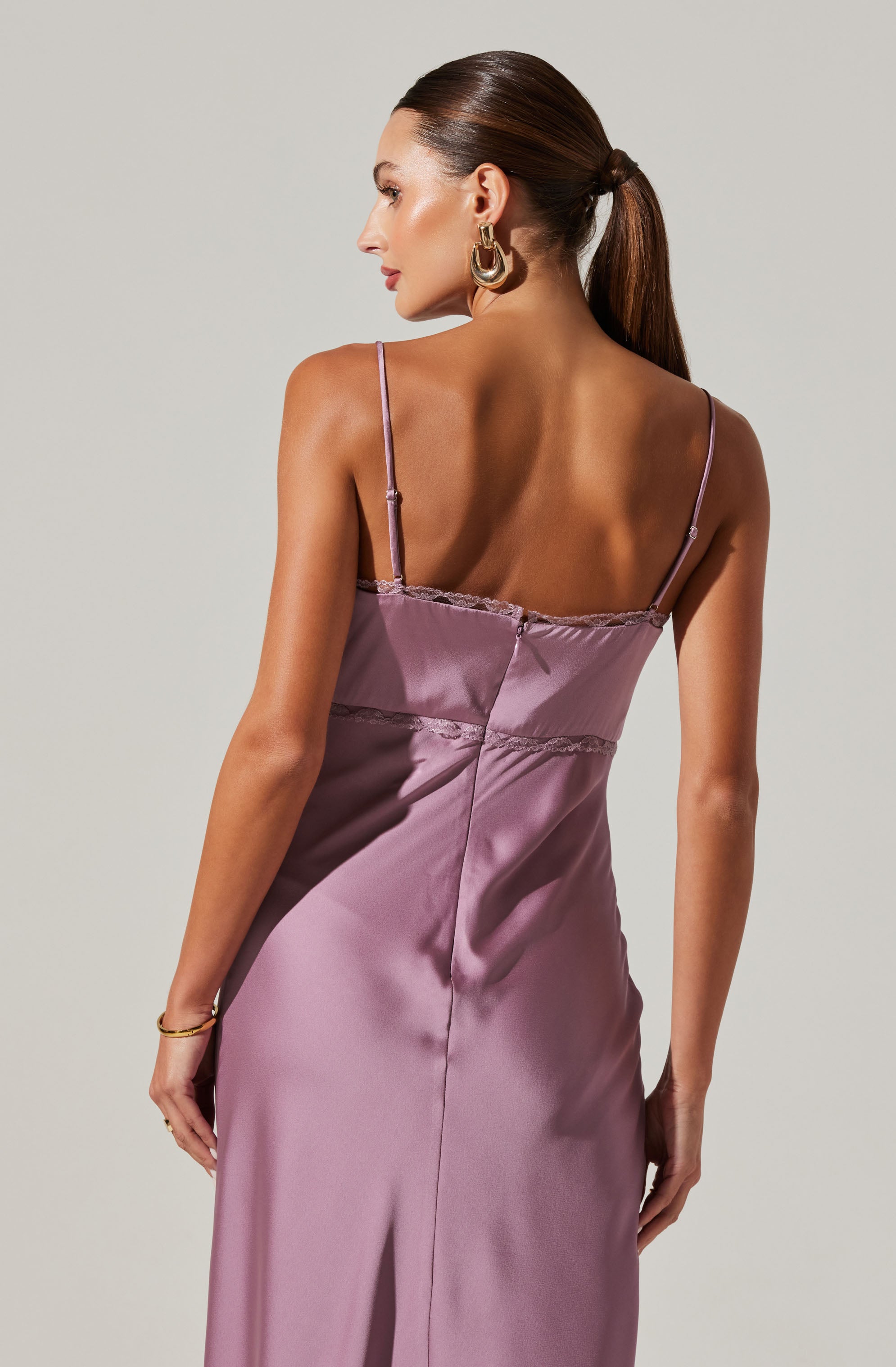 A woman is wearing a silky, mauve-colored slip dress with thin spaghetti straps. The dress features lace detailing along the top edge and a central seam running down the back. She accessorizes with large gold hoop earrings and a delicate bracelet on her right wrist.