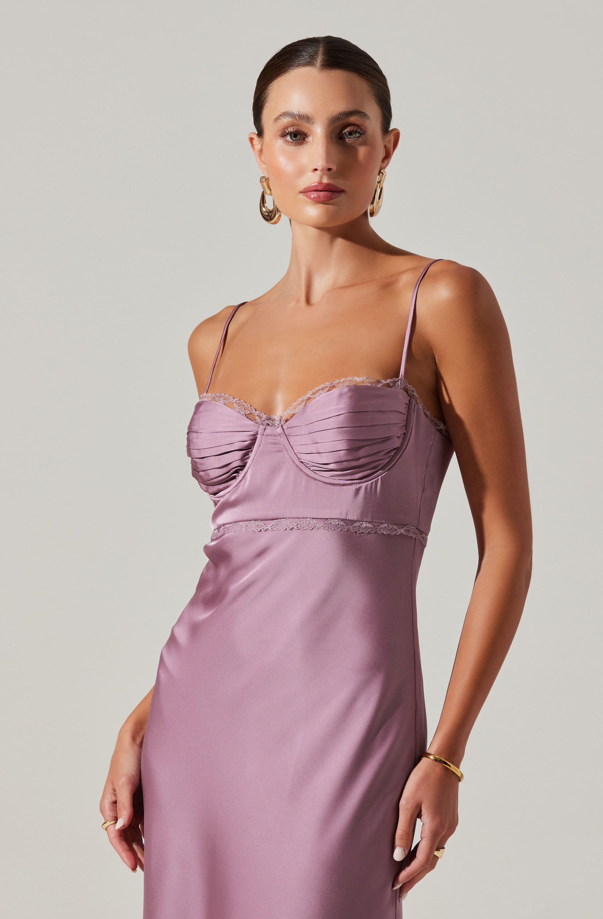 A woman is wearing a silky, lavender-colored slip dress with thin spaghetti straps. The dress features gathered detailing with lace trim along the bust and a smooth, flowing skirt. She has on large, gold hoop earrings and a gold bracelet on her right wrist.