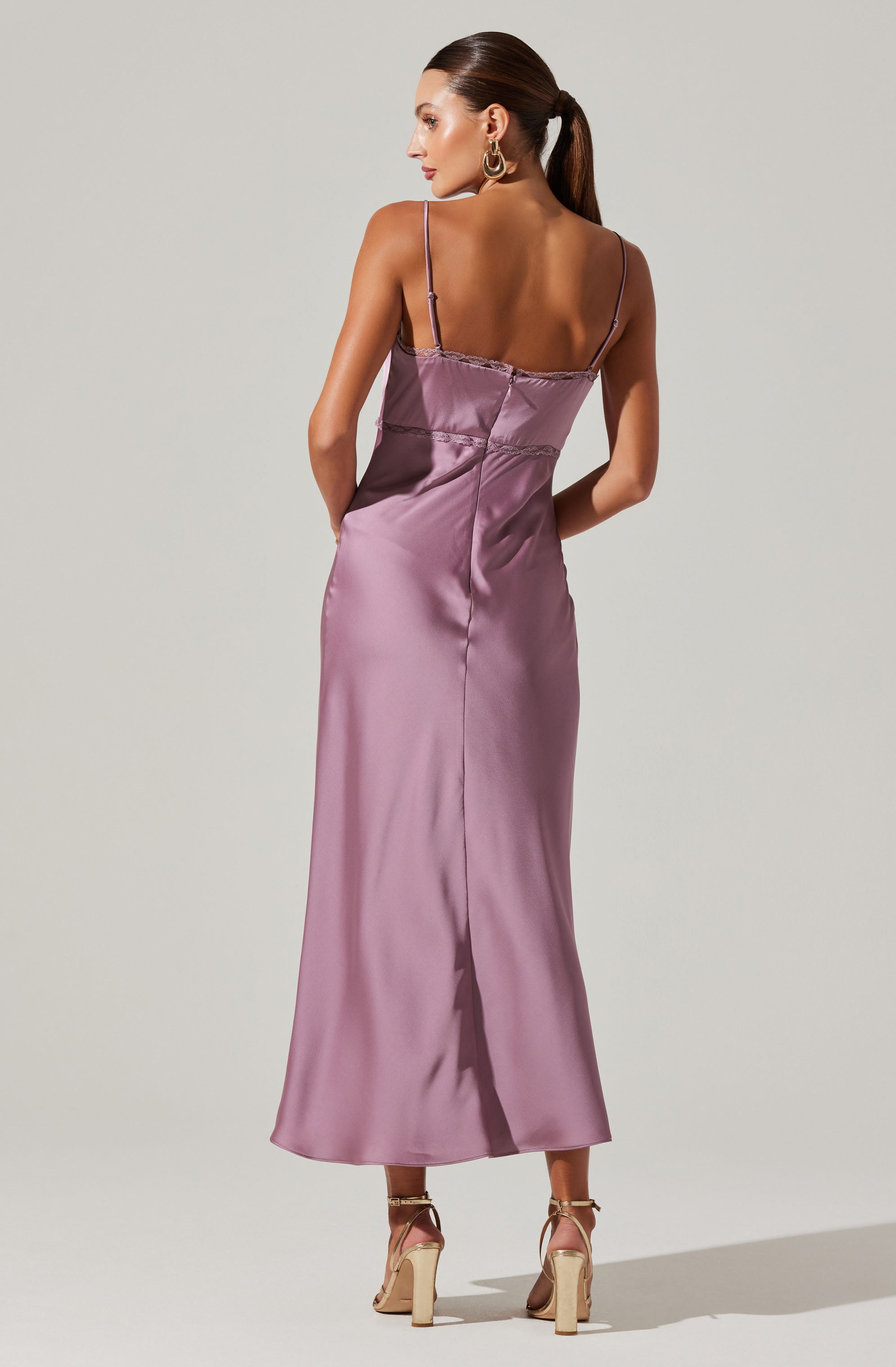 A woman is wearing a long, satin slip dress in a soft lavender color. The dress features thin spaghetti straps and a fitted bodice with a slightly gathered detail at the top. The length extends to the ankles, and the fabric has a smooth, shiny appearance. She is also wearing high-heeled sandals in a neutral shade.