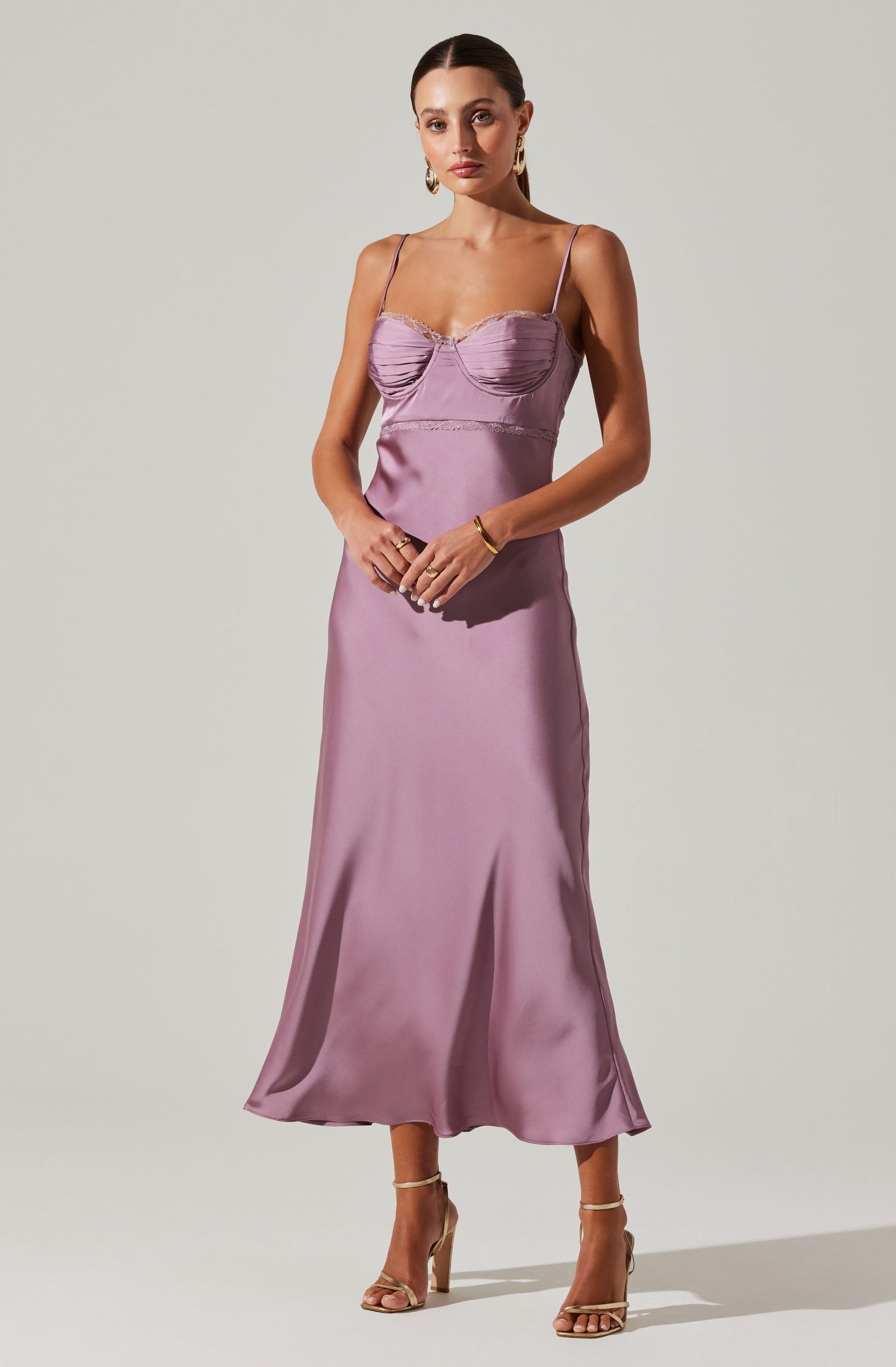 A woman is wearing a lavender satin slip dress with spaghetti straps, a fitted bust with ruching details, and a flowing, ankle-length skirt. She is accessorizing with gold hoop earrings, a gold bracelet, rings, and nude high-heeled sandals.