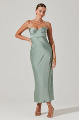A woman is wearing a long, light green satin dress with thin spaghetti straps. The dress features a ruched, fitted bodice with a sweetheart neckline, and the skirt flows straight down to the floor. She accessorizes with gold jewelry, including earrings and a bracelet, and wears beige high-heeled sandals.