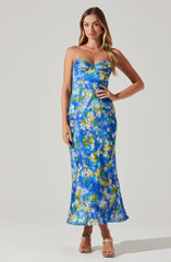 A woman is wearing a long, sleeveless dress with thin spaghetti straps. The dress features a vibrant, abstract floral print in shades of blue, green, yellow, and white. It has a fitted bodice with a sweetheart neckline and a flowy, ankle-length skirt. She is also wearing open-toed, high-heeled sandals with ankle straps.