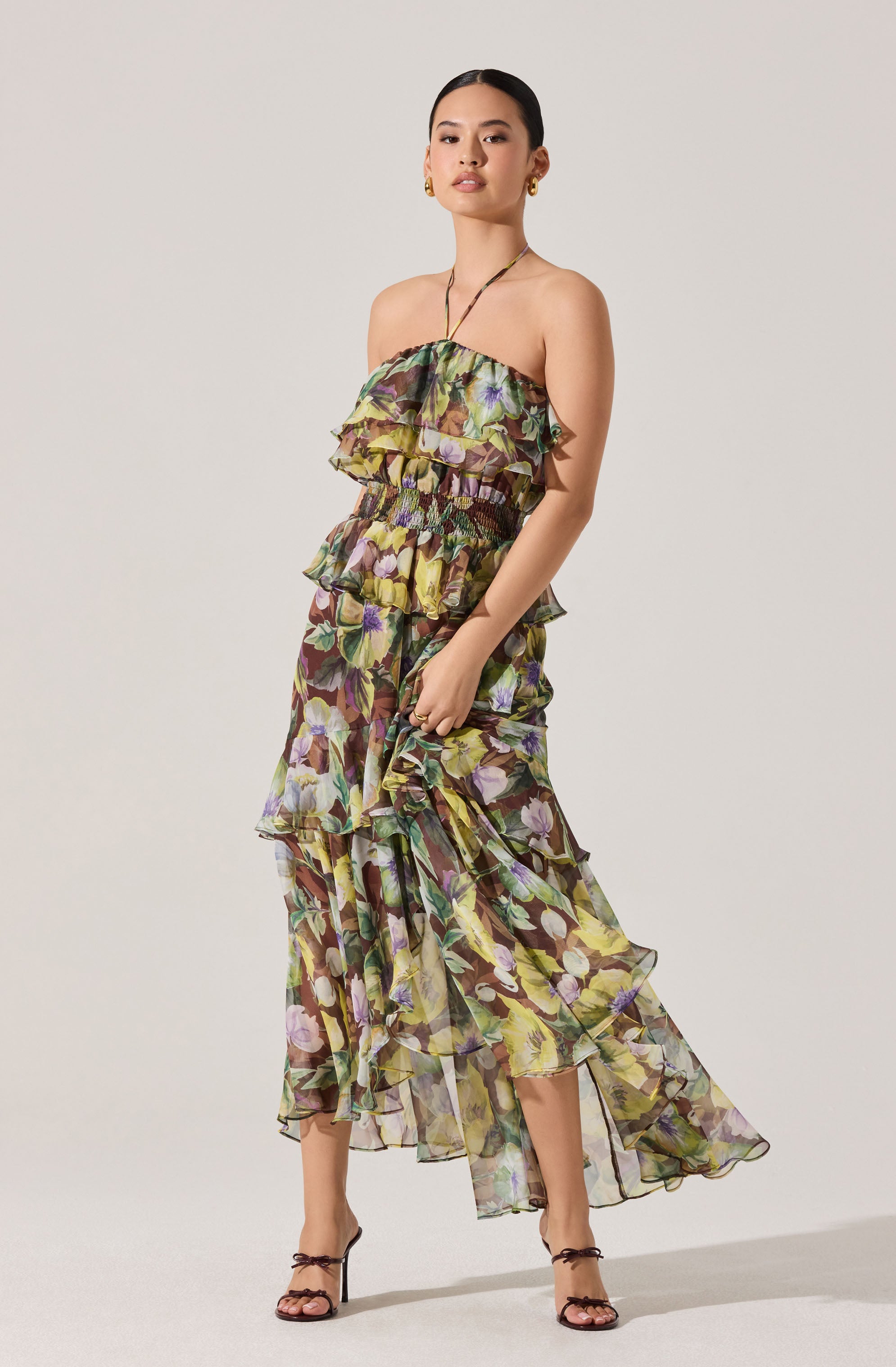 A woman is wearing a strapless, flowy maxi dress with a floral pattern in shades of purple, green, yellow, and brown. The dress features layered ruffles on the bodice and cascading down the length of the skirt. She is also wearing high-heeled sandals with thin ankle straps.