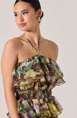 A woman is wearing a sleeveless, strapless dress with a floral pattern featuring shades of green, yellow, purple, and brown. The dress has layered ruffles and a smocked waistband. She accessorizes with large gold hoop earrings and a delicate necklace with a thin chain. Her hair is pulled back into a sleek, low bun.