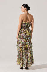 A woman is wearing a long, sleeveless dress with a floral pattern. The dress features a halter neck with a tie at the back, a ruffled top, and multiple tiers of ruffles along the skirt. The fabric is sheer and lightweight with colors including shades of purple, green, yellow, and brown.