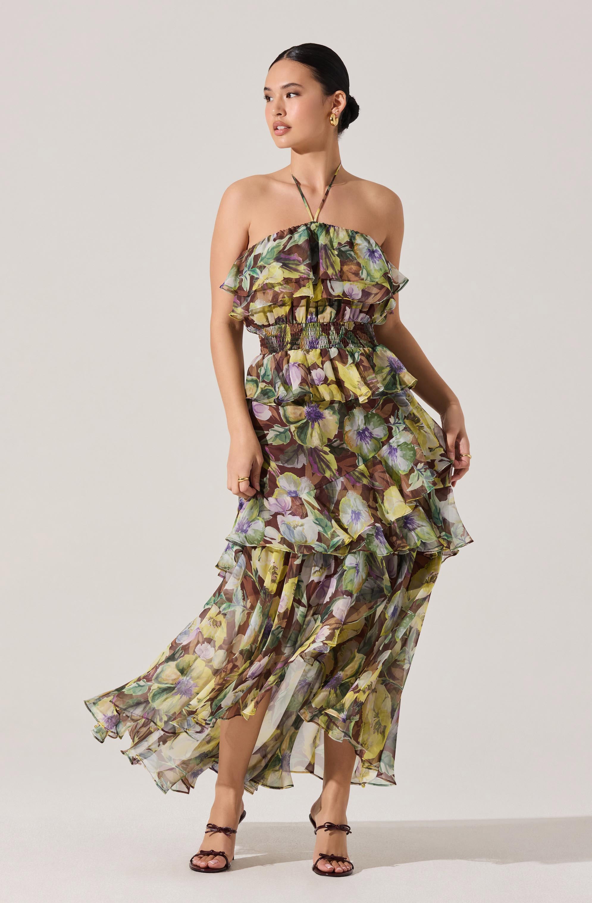A woman is wearing a strapless, flowy dress with a layered, ruffled design. The dress features a colorful floral pattern with shades of purple, yellow, green, and brown. The fabric is lightweight and semi-sheer, giving a breezy, elegant appearance. She is also wearing open-toed black high-heeled sandals with ankle straps.