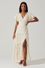 A woman is wearing an off-white, floor-length dress with a deep V-neckline and short, ruffled sleeves. The dress features a high waist, tiered skirt with a slit up the front, and subtle embroidered floral details throughout. She has accessorized with gold earrings, a bracelet, and strappy high heels.