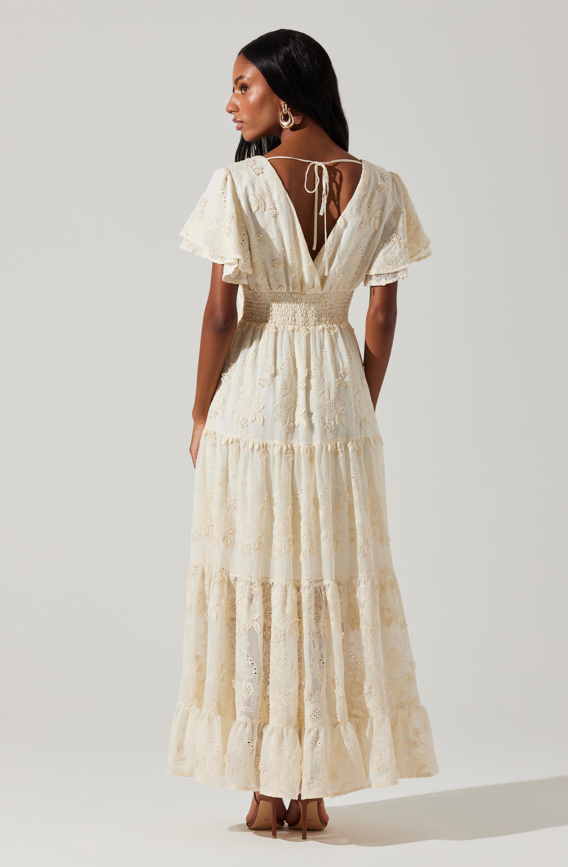 A woman is wearing a long, cream-colored dress with eyelet embroidery. The dress features short, flutter sleeves and a deep V back with a tie closure at the neckline. The waistline is fitted with a smocked detail, and the skirt has a tiered design with multiple gathered sections, creating a flowing and layered appearance. She is also wearing large gold hoop earrings and beige high-heeled shoes.