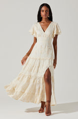 A woman is wearing a long, cream-colored lace dress with a V-neckline and short, ruffled sleeves. The dress has a fitted waist and a tiered skirt with a high slit on the left side, revealing part of her leg. She is accessorized with bracelets, rings, and dangling earrings, and is wearing strappy high-heeled sandals.