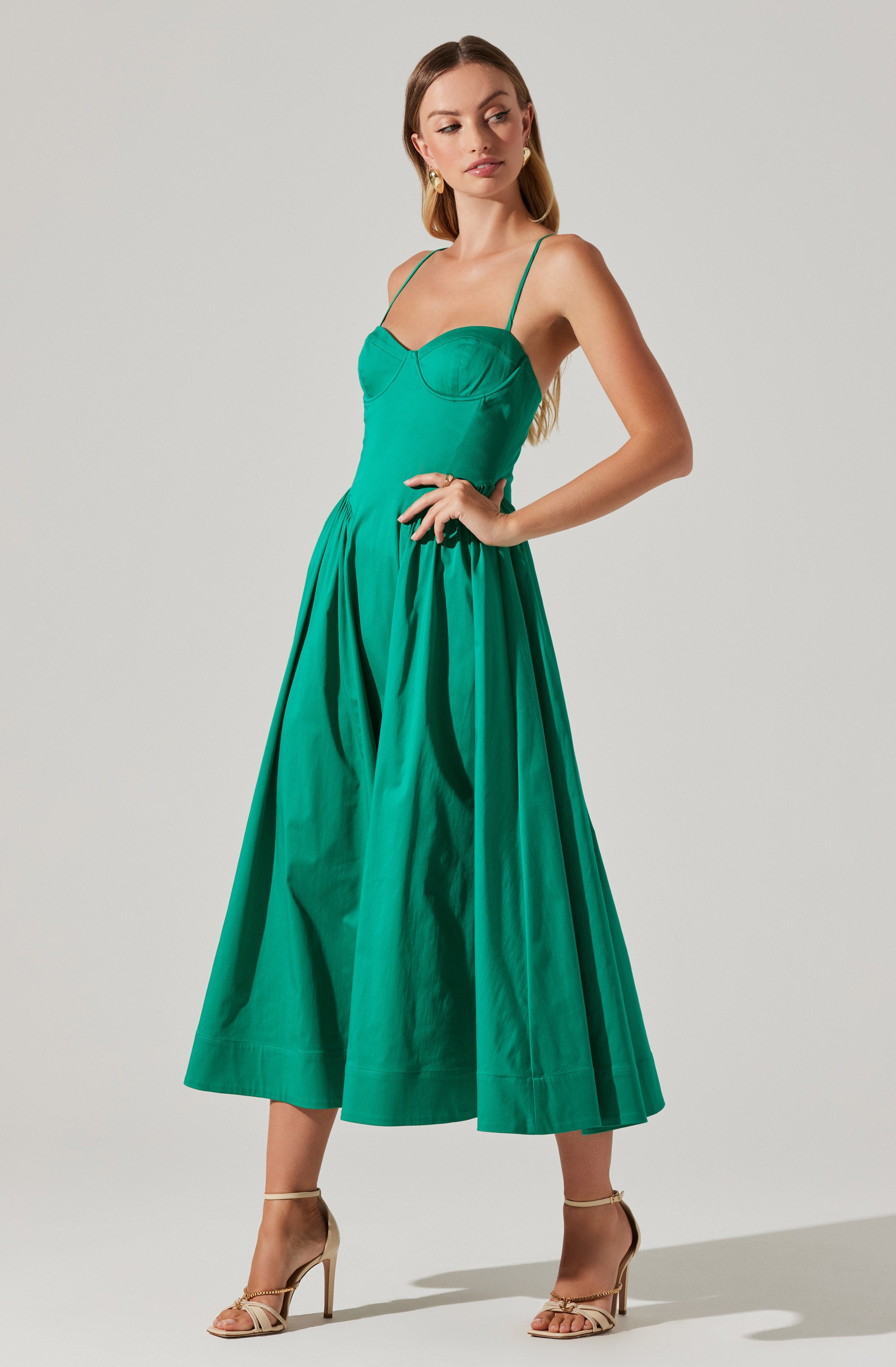 A woman is wearing a sleeveless, green dress with thin spaghetti straps and a fitted bodice that features a sweetheart neckline. The dress has a high waistline and a full, pleated skirt that falls to her mid-calf. She is accessorized with gold drop earrings and beige high-heeled sandals.
