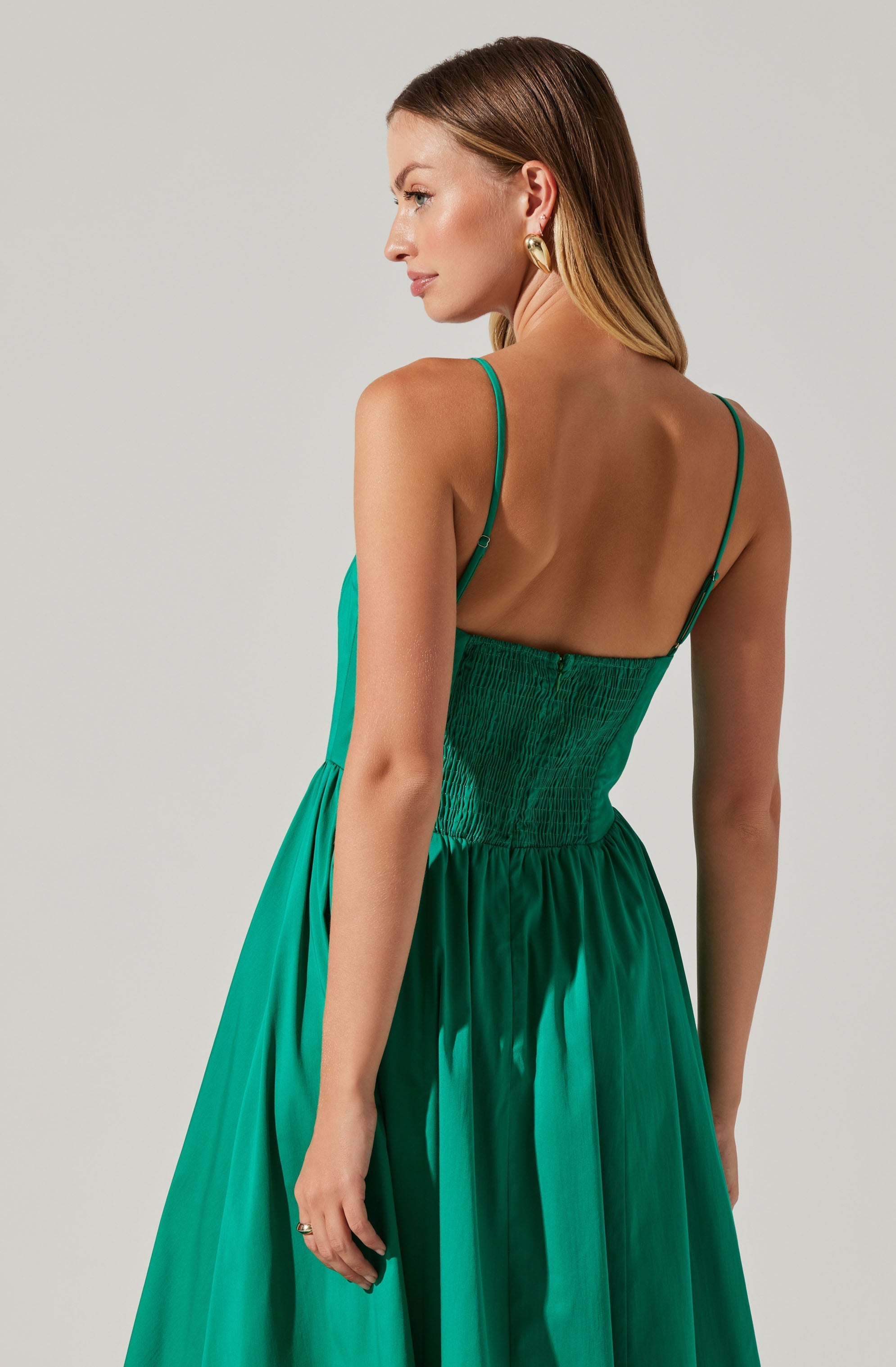 A woman is wearing a sleeveless, emerald green dress with thin spaghetti straps. The dress features a fitted, shirred bodice and a full, pleated skirt. She has light brown hair styled straight, and she is wearing large, gold hoop earrings.