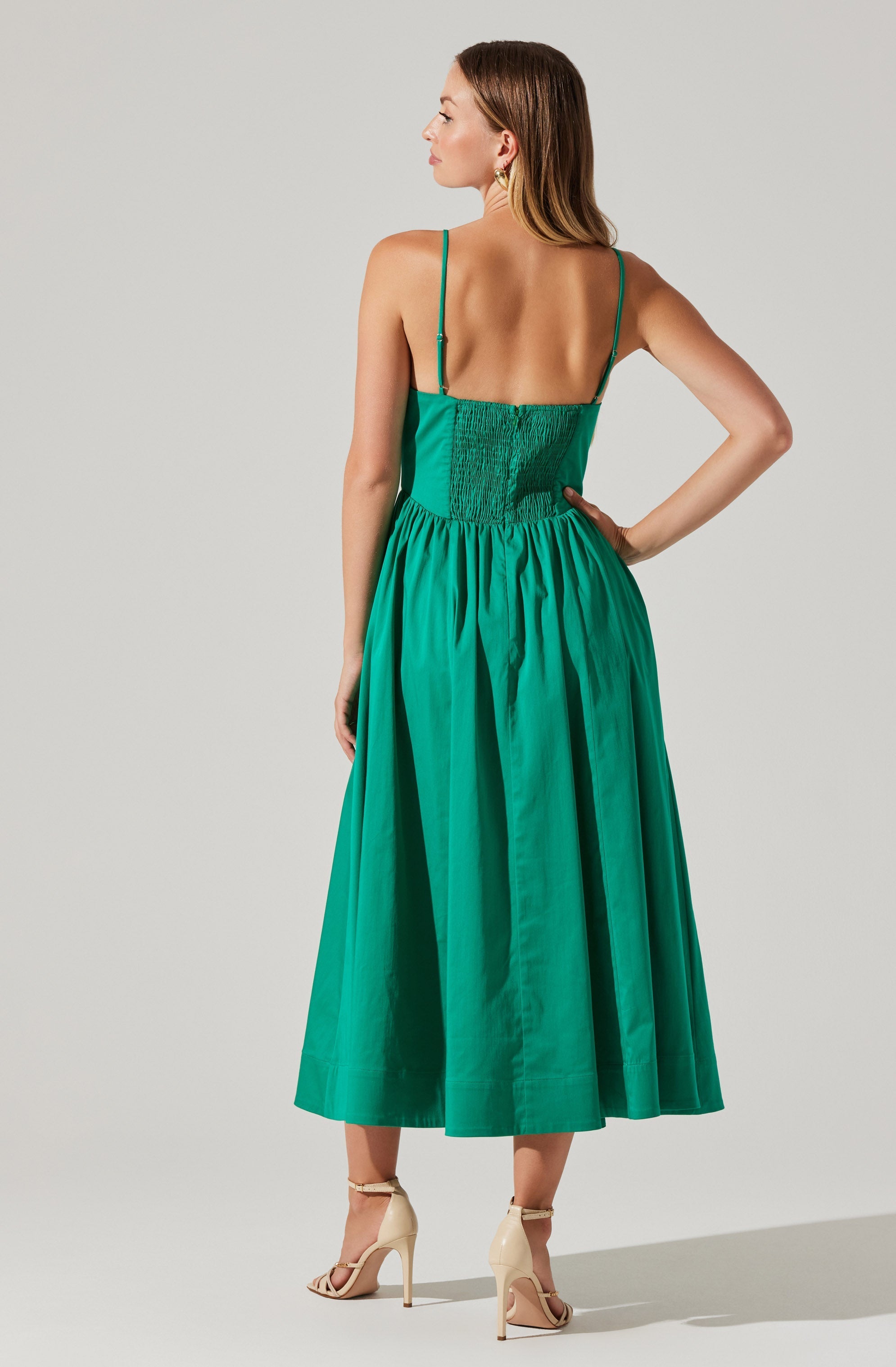 A woman is wearing a sleeveless, backless midi dress with thin spaghetti straps. The dress features a fitted, smocked bodice and a full, gathered skirt made of vibrant green fabric. She is also wearing nude high-heeled sandals with ankle straps.