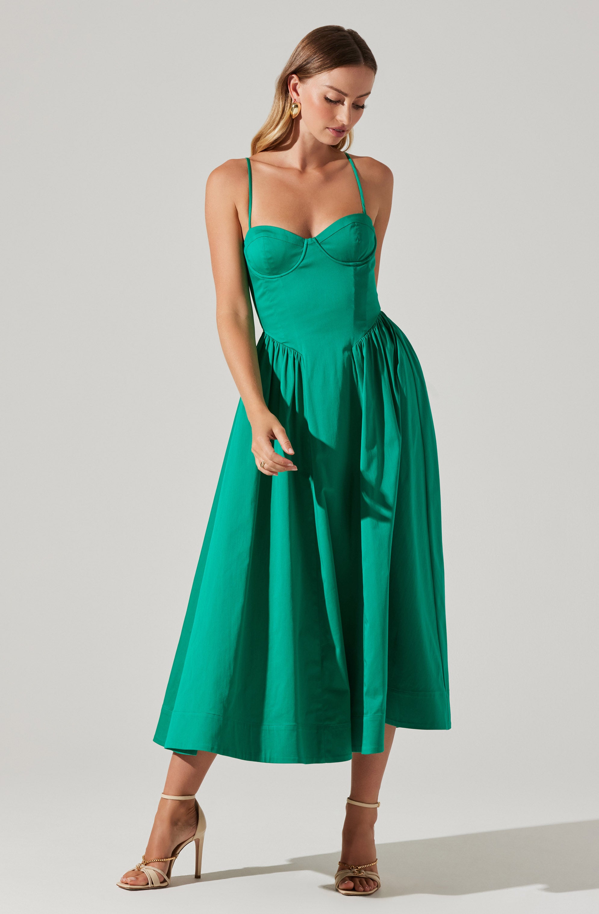 A woman is wearing a long, sleeveless dress with thin spaghetti straps. The dress has a fitted bodice with a structured top and a full, flowing skirt that extends to her ankles. The fabric appears shiny and smooth, and the dress is a vibrant shade of teal. She is also wearing high-heeled sandals in a neutral color.