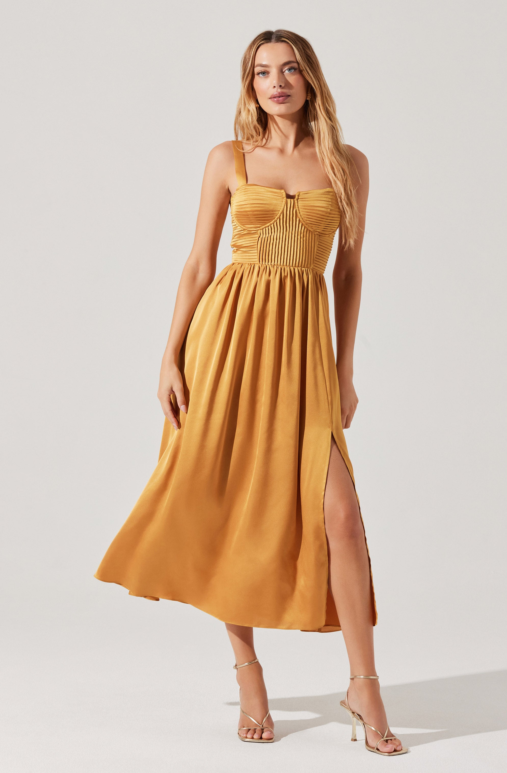 A woman is wearing a sleeveless, mustard-yellow dress with thin spaghetti straps. The bodice features horizontal and vertical pleats and a small keyhole opening in the center. The dress has a fitted waist and a full, gathered skirt that reaches the ankles, with a high slit on one side revealing her leg. She is also wearing strappy high-heeled sandals.