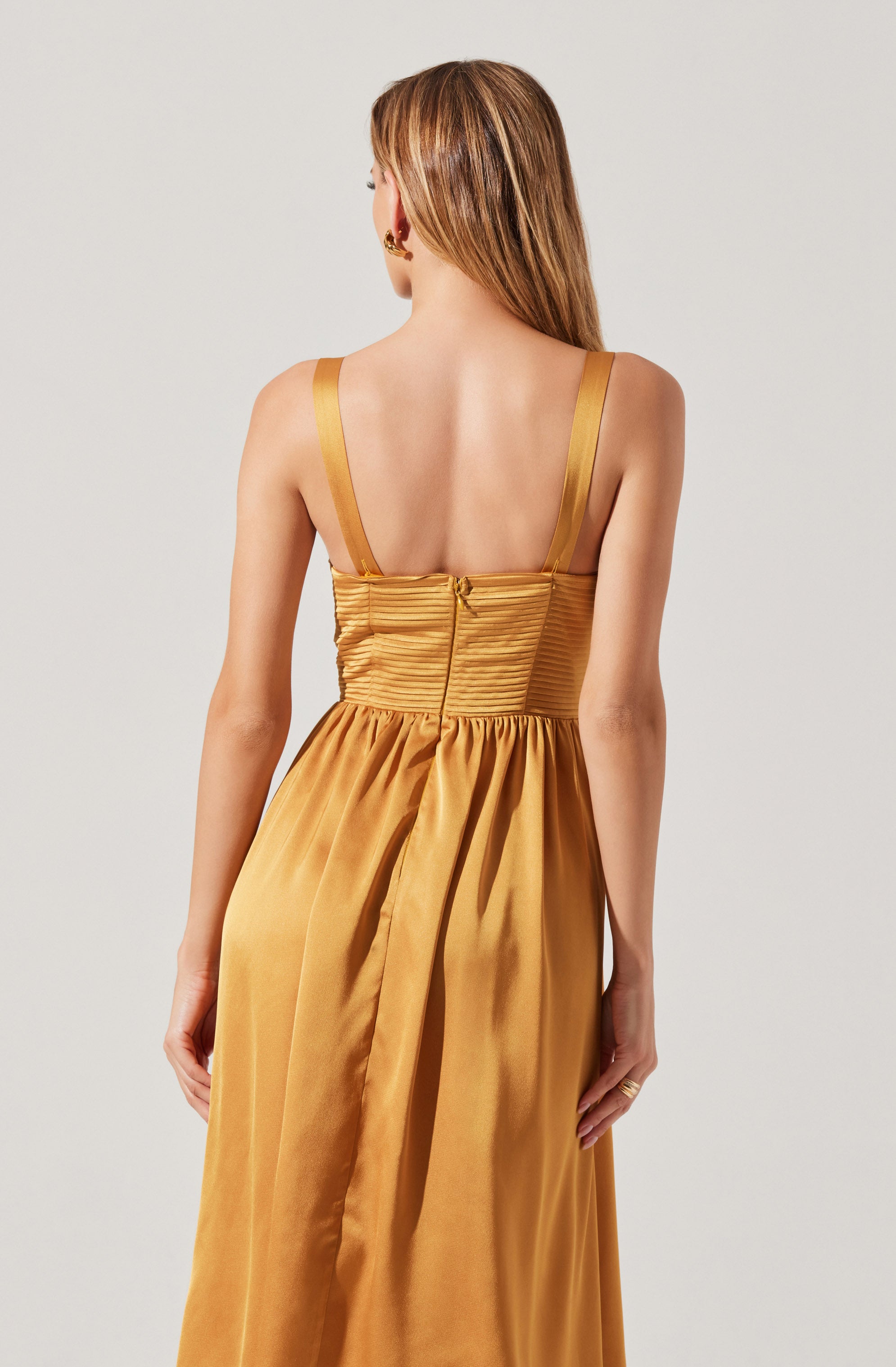 The dress has wide, shiny straps over the shoulders and a fitted, ruched bodice that is also shiny and pleated. The fabric appears to be satin or silk, and it is a warm golden color. The back of the dress features a zipper closure and a full, flowing skirt that extends to the floor.