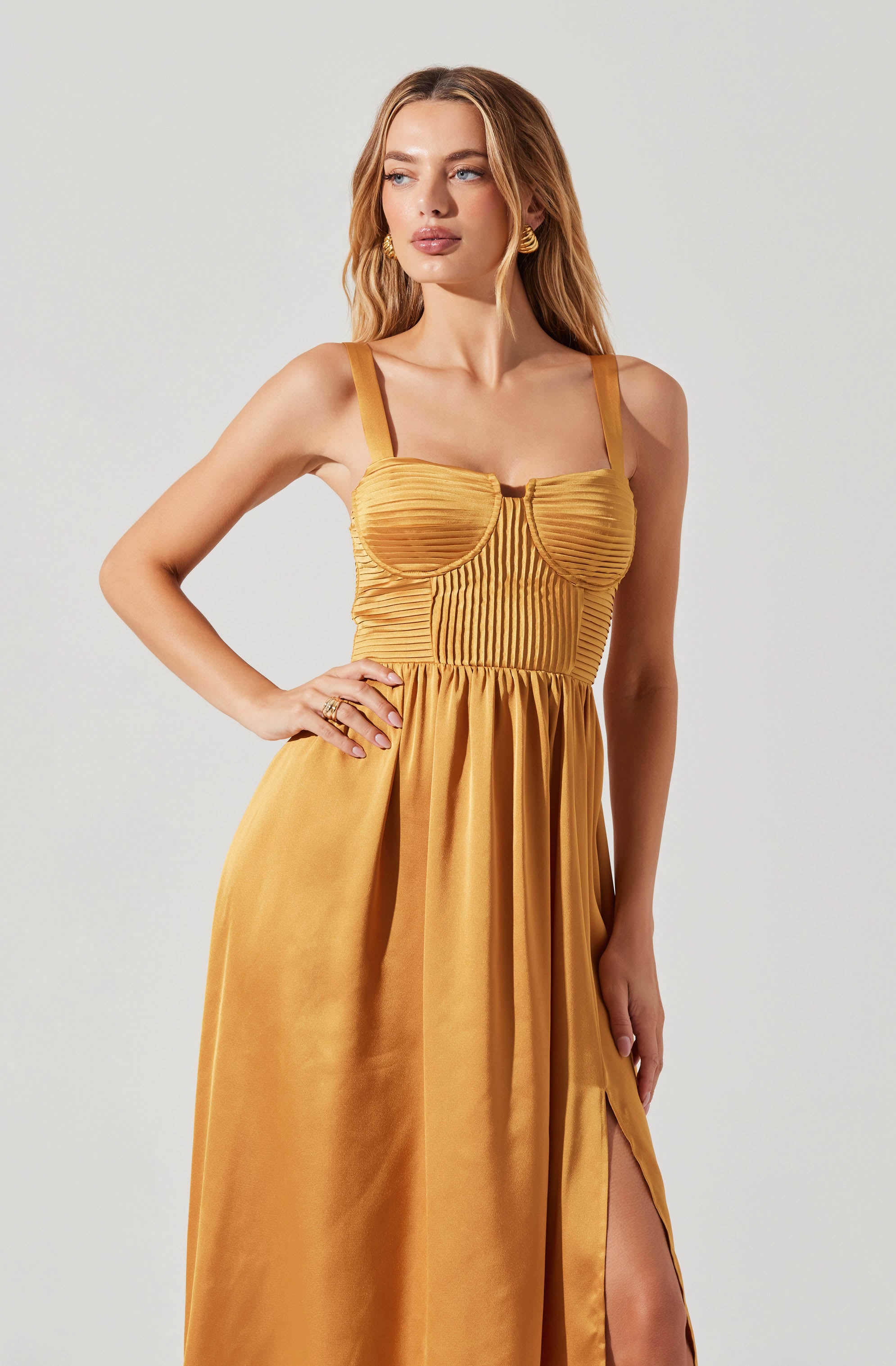 A woman is wearing a sleeveless, mustard yellow dress with wide shoulder straps. The bodice features a structured, ribbed design with vertical pleats and a small cutout at the center neckline. The skirt is flowing, gathered at the waist, and has a high slit on the side.