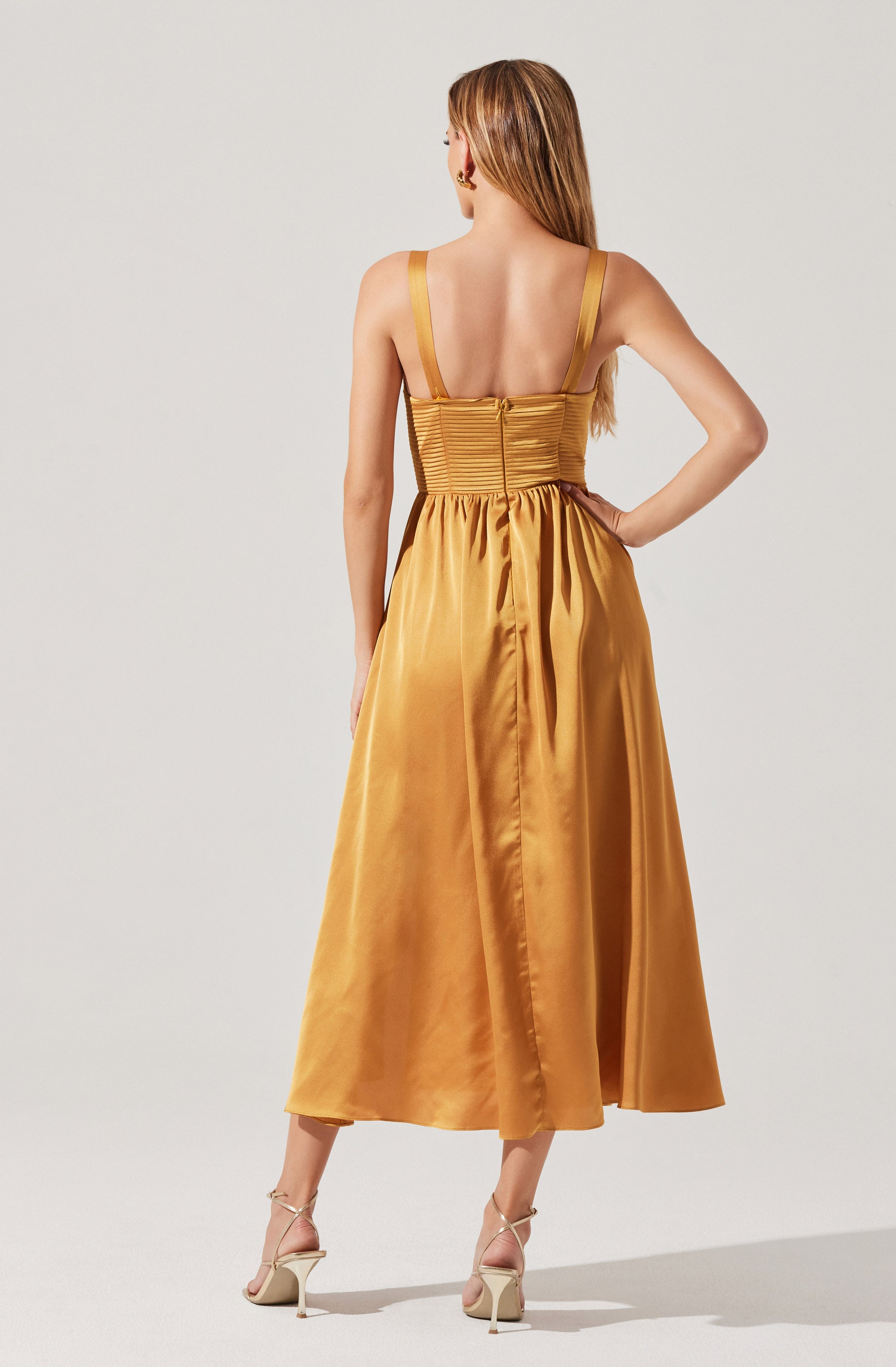 A woman is wearing a sleeveless, gold satin dress with wide shoulder straps, a fitted, ruched bodice, and a full-length skirt. The dress has a back zipper closure and a smooth, shiny texture. She is also wearing beige high-heeled sandals.