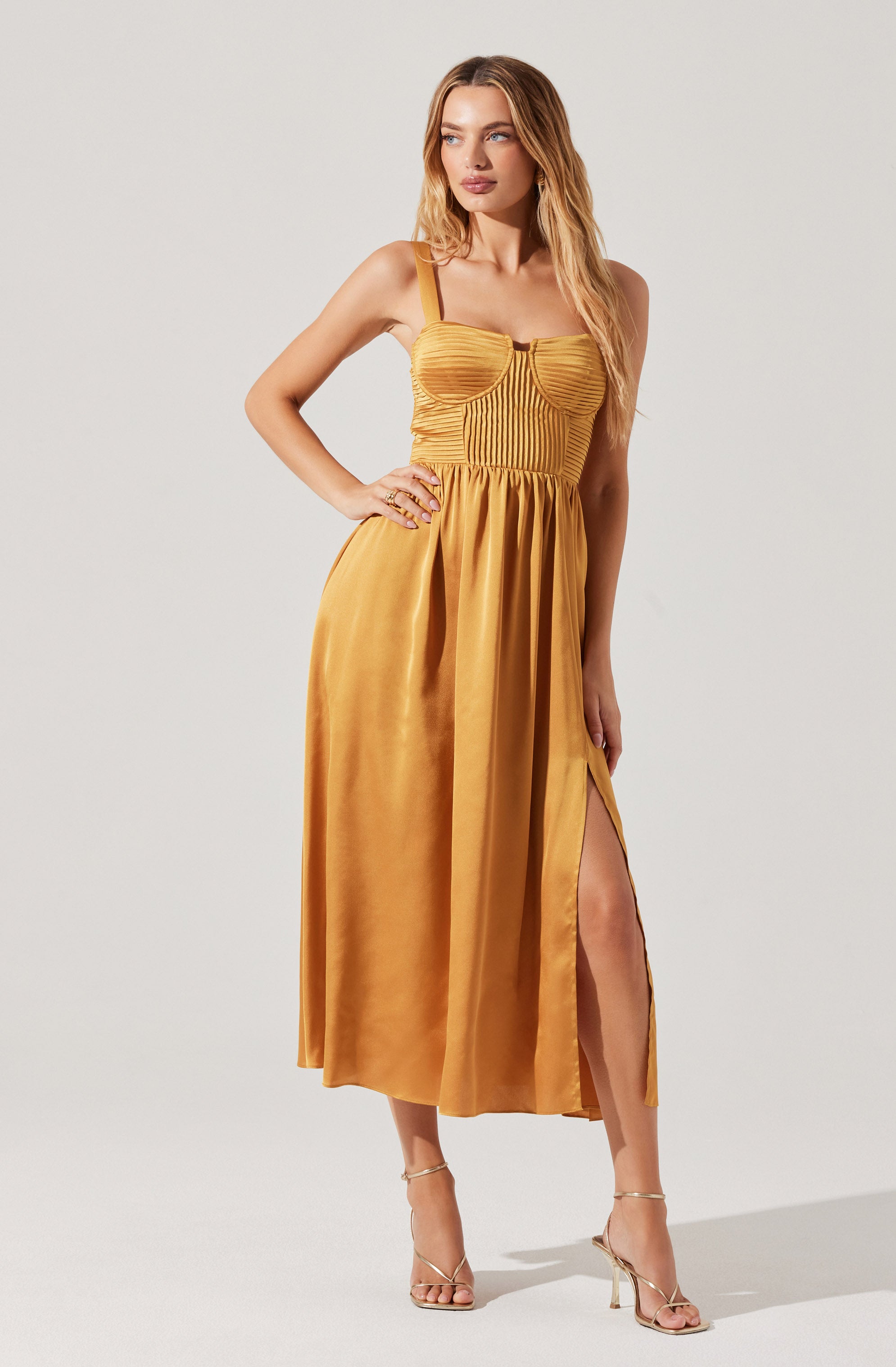 A woman is wearing a sleeveless, mustard yellow dress with a fitted, ruched bodice and spaghetti straps. The dress has a high slit on the right side, revealing her leg. She is also wearing strappy beige high-heeled sandals.